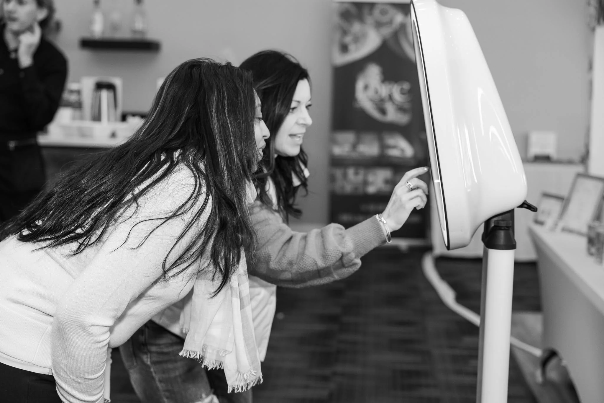 Two women are leaning forward and looking at a monitor in a modern office or classroom setting.