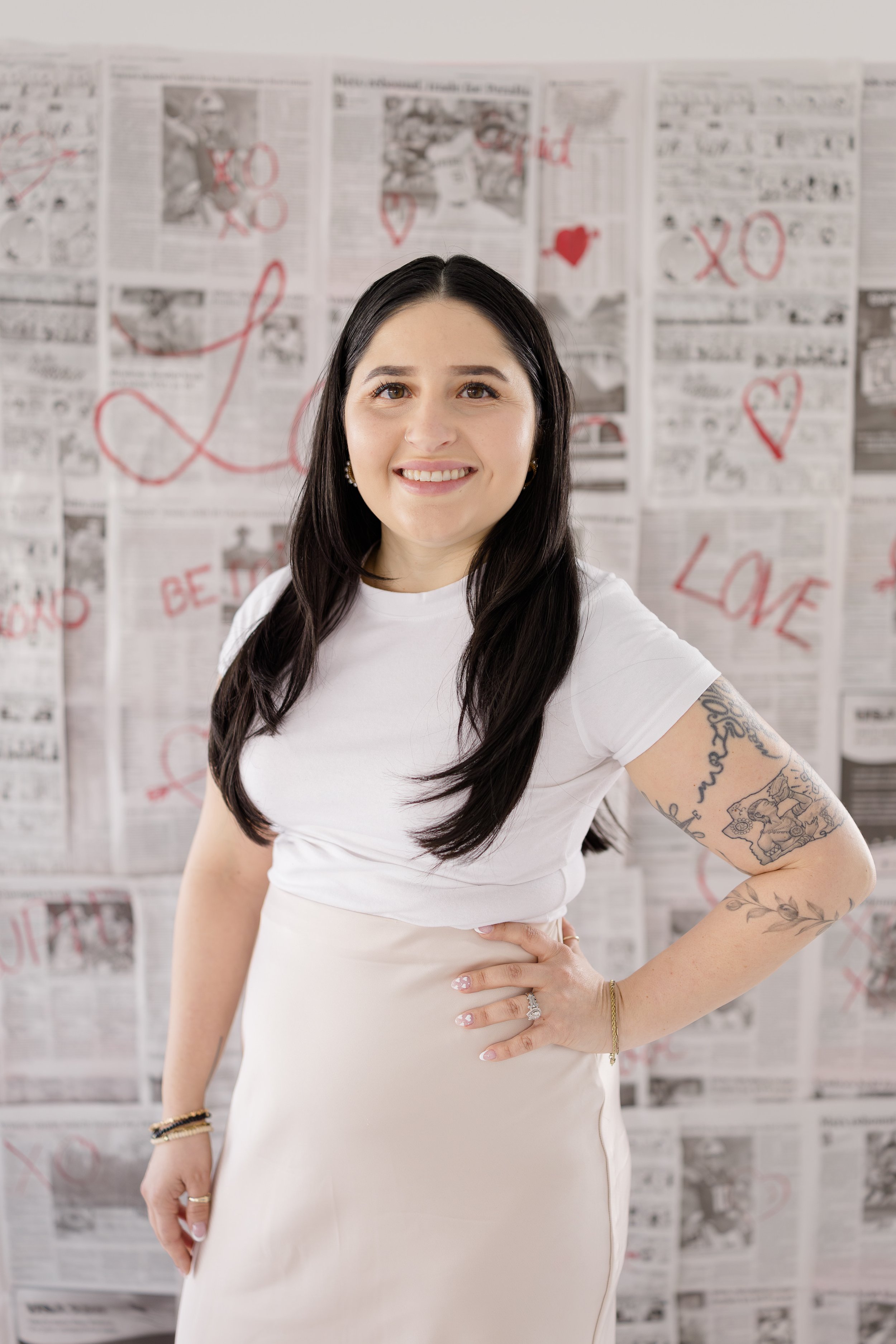 A woman with long black hair, wearing a white short-sleeved top and a beige skirt, standing in front of a wall decorated with newspaper pages with red heart and love-themed drawings and writings, smiling at the camera.
