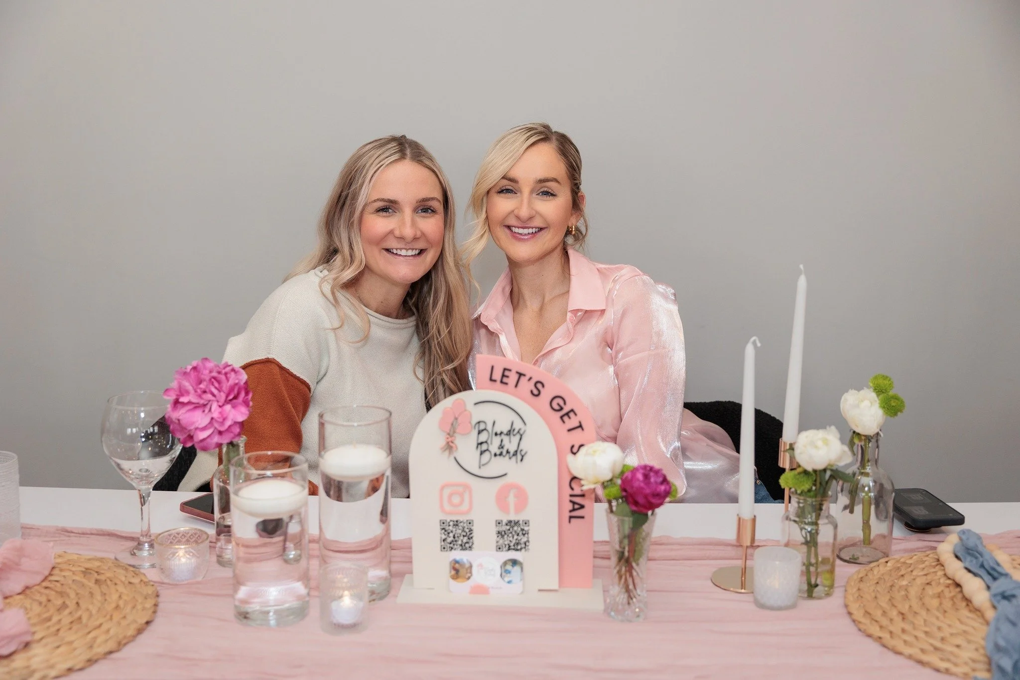 Two women smiling at a Hello Kitty-themed birthday party table decorated with pink and white flowers, candles, and drinks.