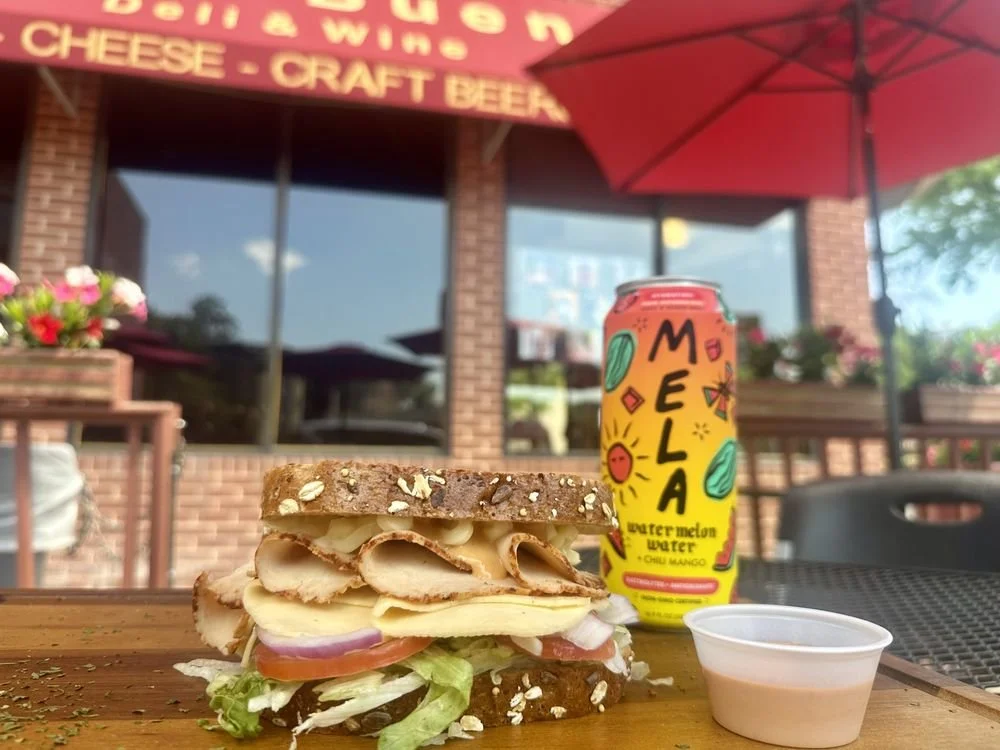 A sandwich on a wooden table with layers of sliced turkey, cheese, tomato, onion, and lettuce, served with a side of sauce in a small cup, a can of Mela sparkling water, and a pink umbrella in the background outside a restaurant with a brick exterior and a red awning.