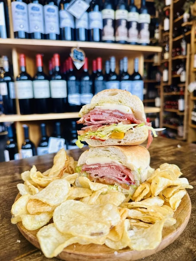 Stacked sandwich with ham, cheese, lettuce, tomato, and pickles on a bun, served with potato chips on a wooden plate, in front of wine bottles on wooden shelves.