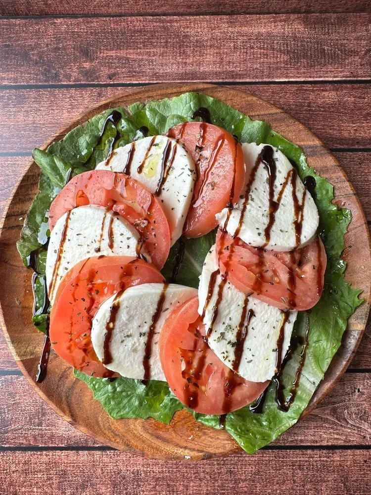Caprese salad with sliced mozzarella cheese, tomato slices, basil leaves, drizzled with balsamic glaze and olive oil, served on a wooden plate with a bed of lettuce.