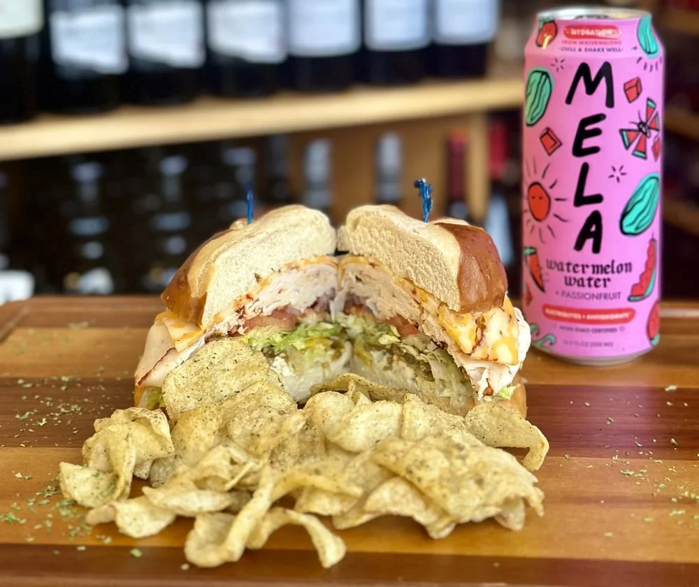 Half of a sandwich with lettuce, tomato, turkey, cheese, and mayonnaise, served with potato chips and a can of watermelon flavored sparkling water on a wooden table.