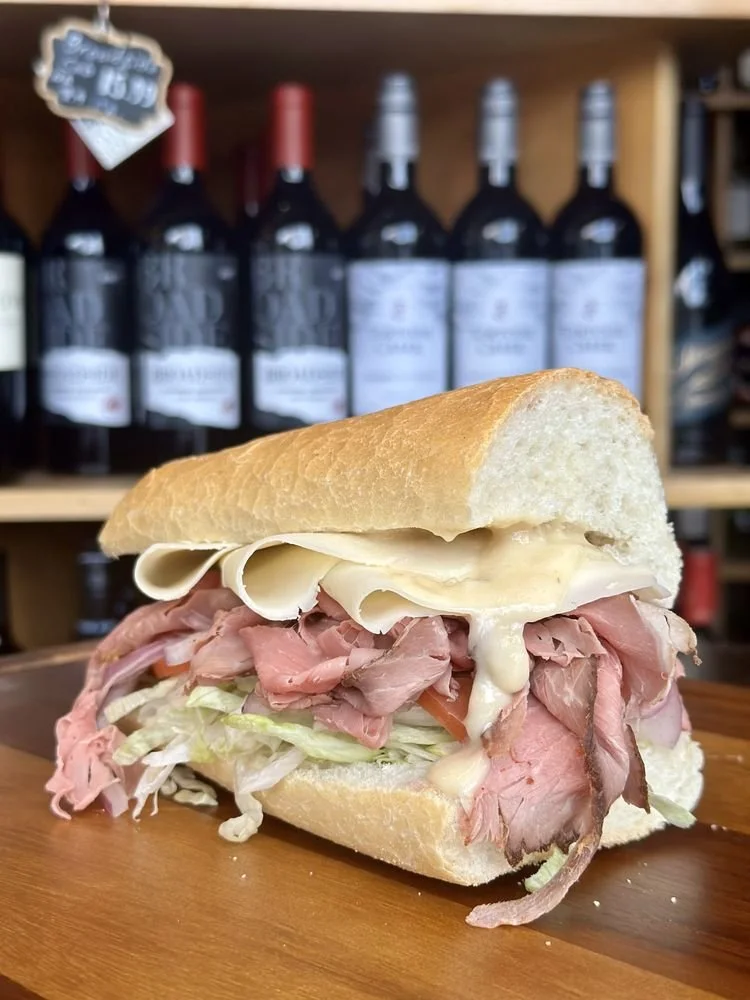 A sandwich with roast beef, lettuce, tomato, and cheese on a sub roll, placed on a wooden surface with bottles of wine in the background.