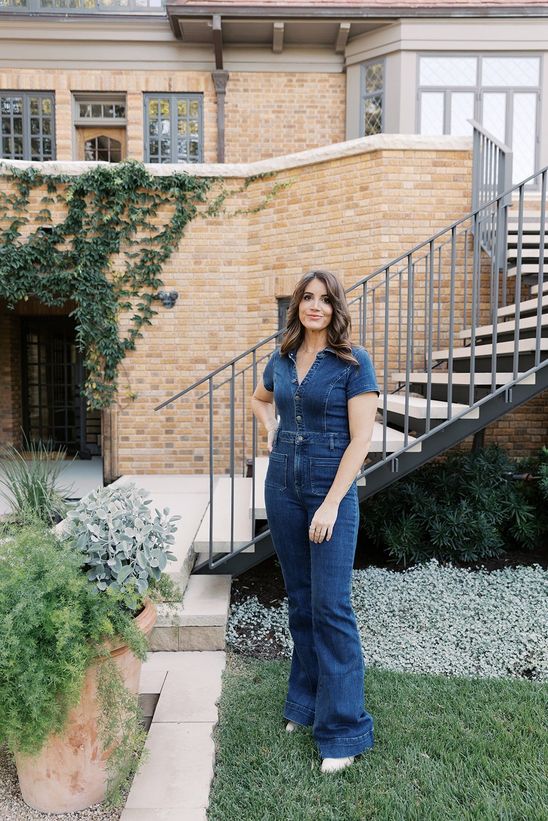 A woman wearing a denim jumpsuit standing outdoors in front of a brick house with garden plants and stairs.