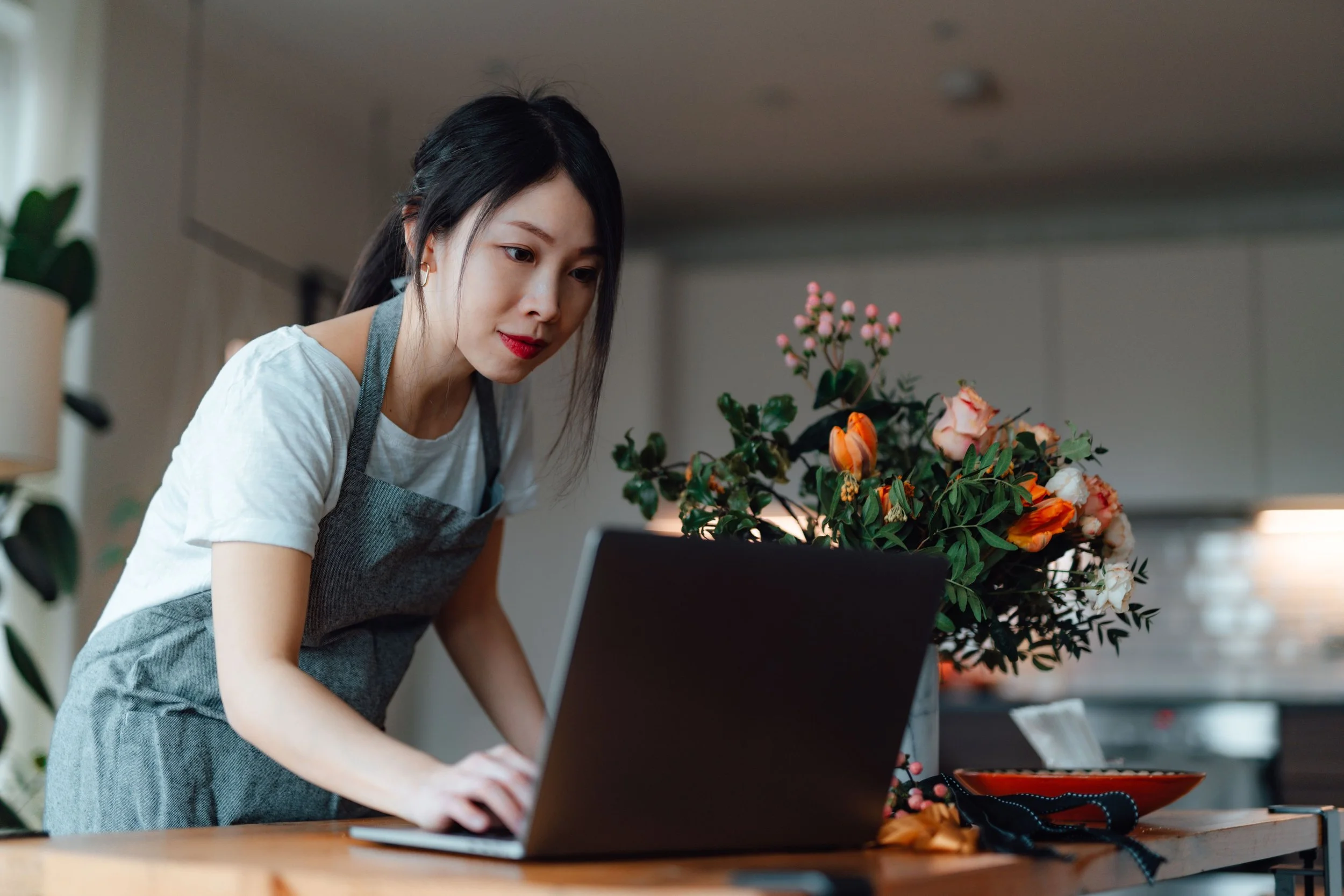 A woman with dark hair in a ponytail working on a laptop at a wooden table, wearing a gray apron over a white t-shirt, with flowers and plants in the background.