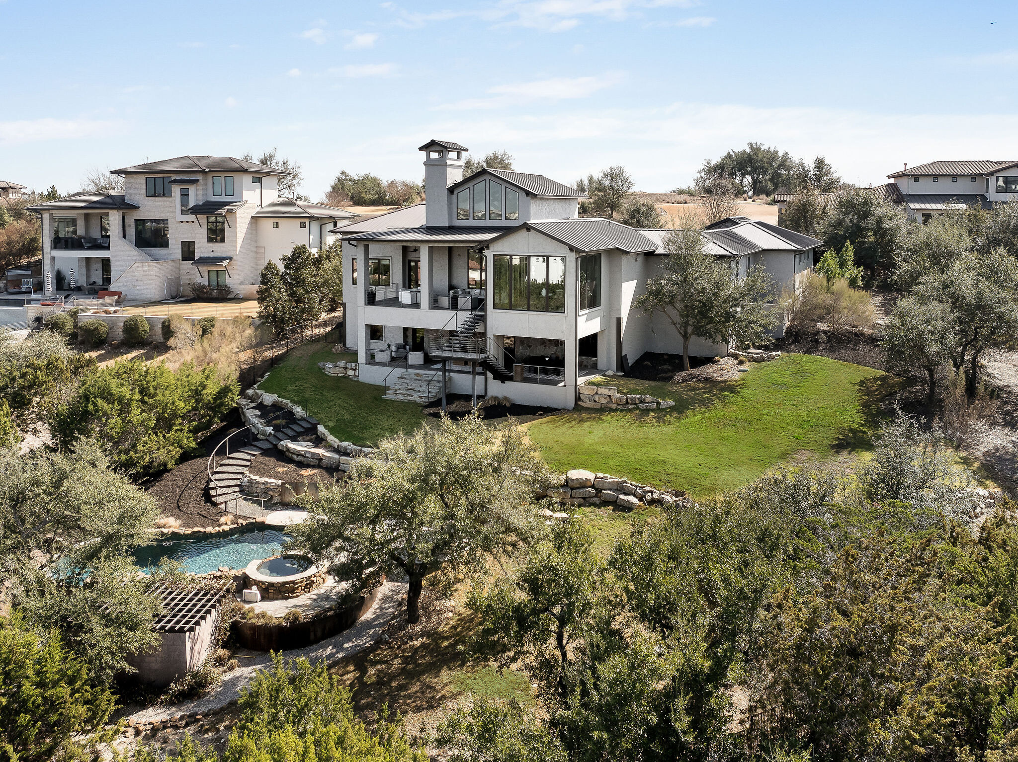 Sweeping rear exterior of the estate at 11928 Musket Rim in Spanish Oaks, showcasing terraced entertaining decks, a dramatic pool grotto with waterfall and heated spa, and panoramic Hill Country views from a commanding 1.36-acre hillside setting.