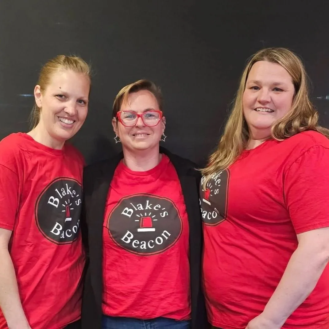 Three women standing together wearing matching red t-shirts with a logo that reads 'Blake's Beacon' and an image of a beacon light. They are smiling and posing against a dark background.