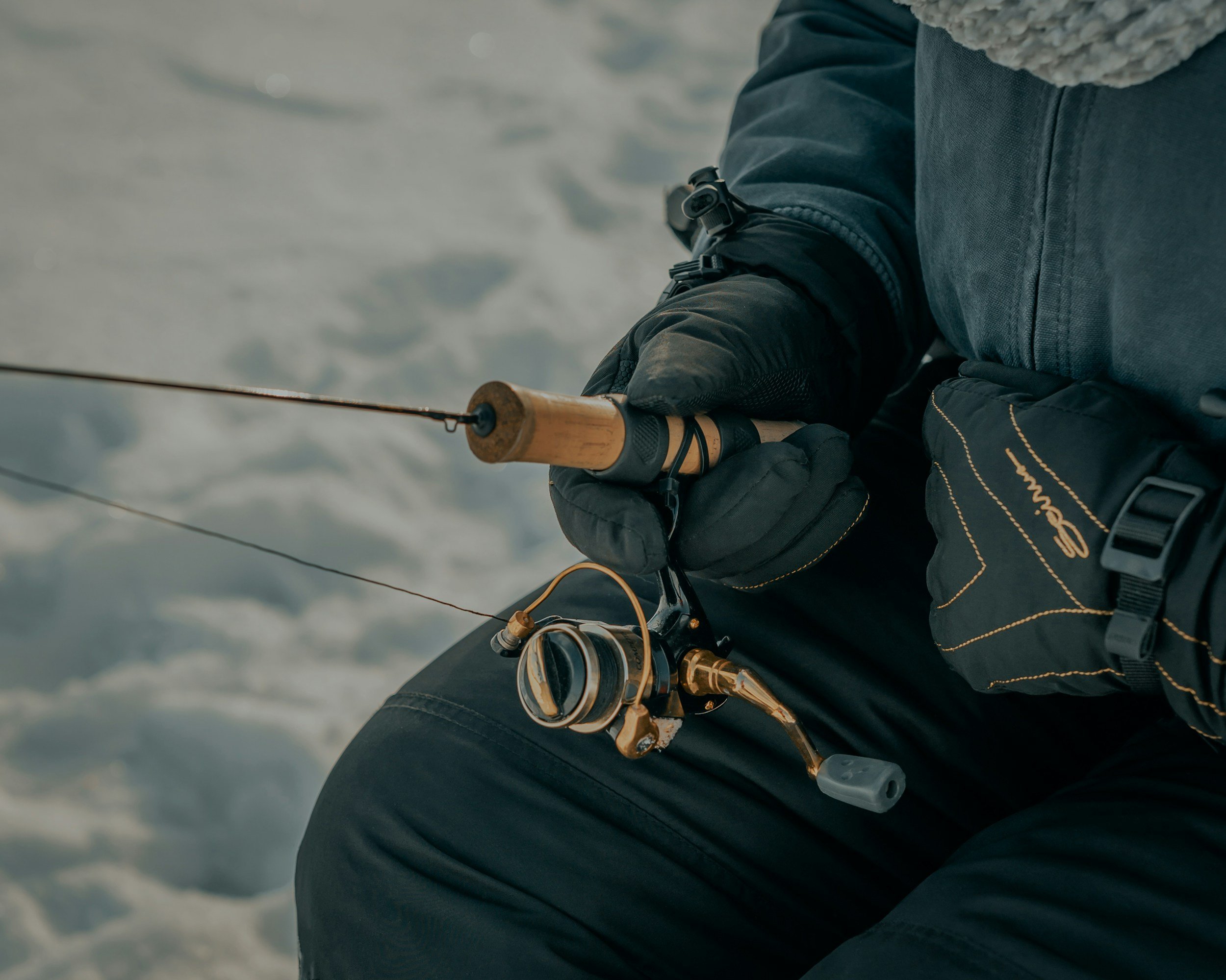 Close-up of a person fishing with a spinning rod and reel, wearing black gloves and jeans, with snow or ice in the background.