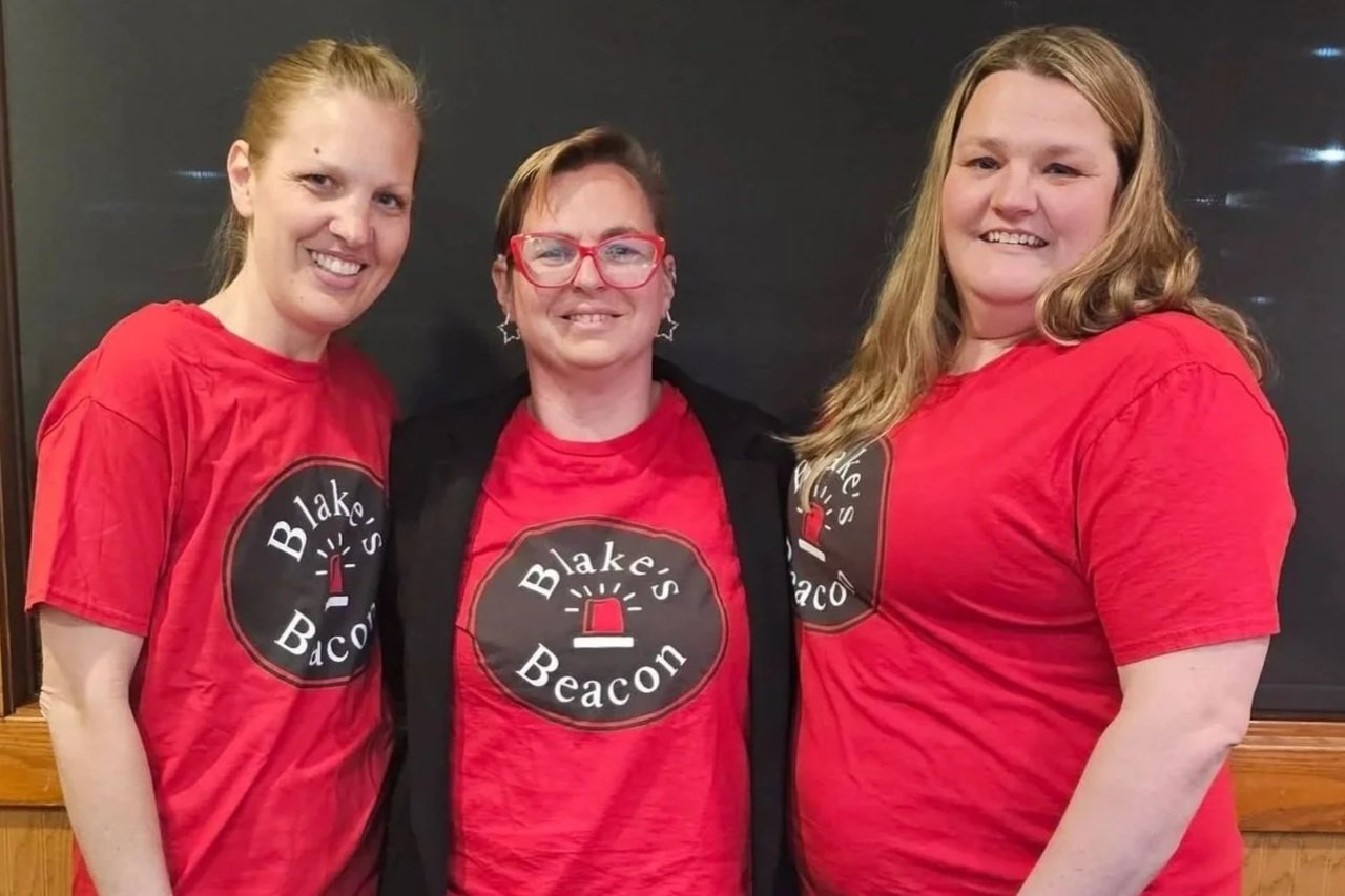 Three women smiling, wearing red T-shirts with a logo that reads 'Blake's Beacon' and features a red emergency siren icon.