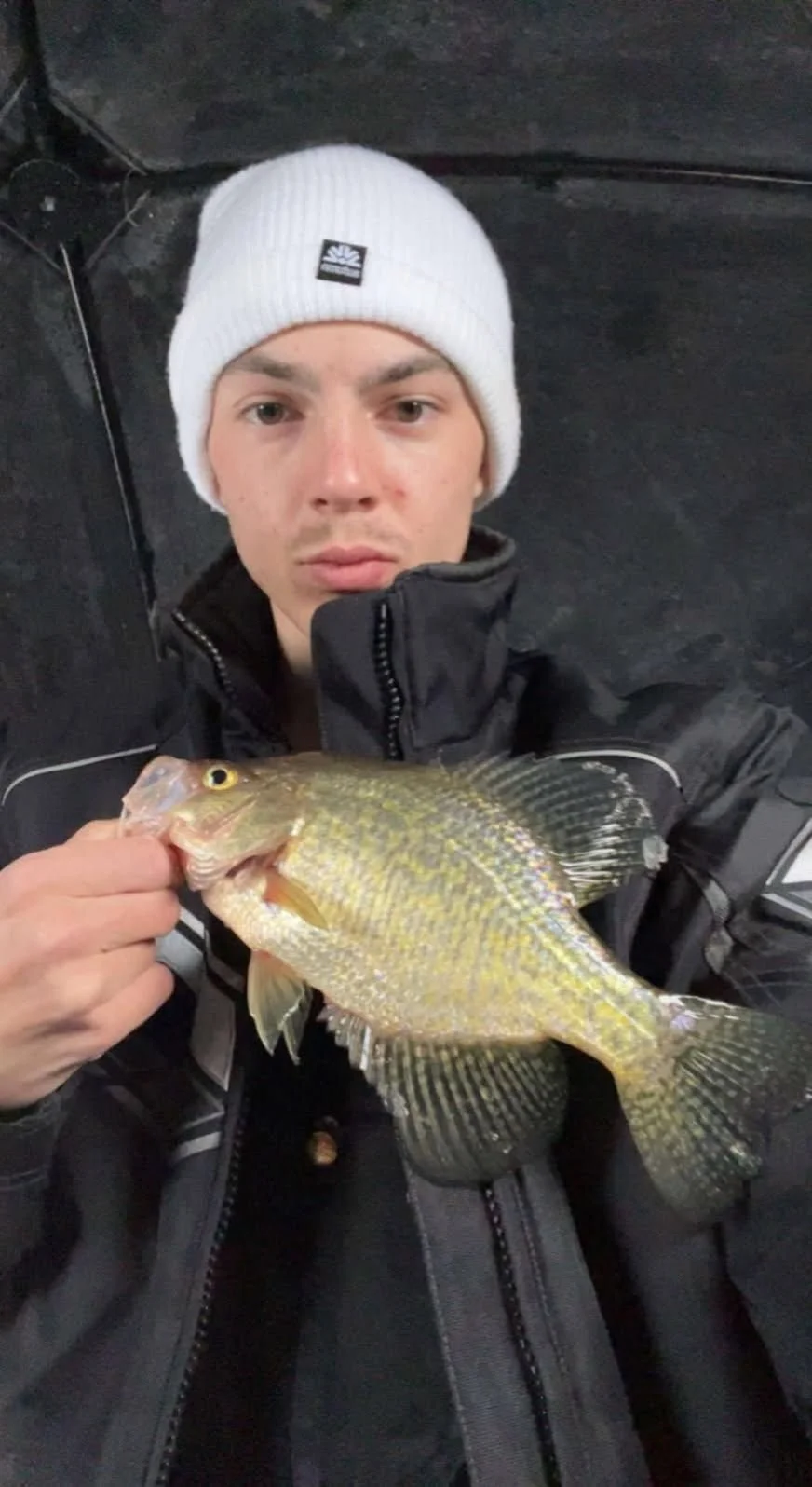Young man in a white beanie and black jacket holding a fish with yellowish-green scales.