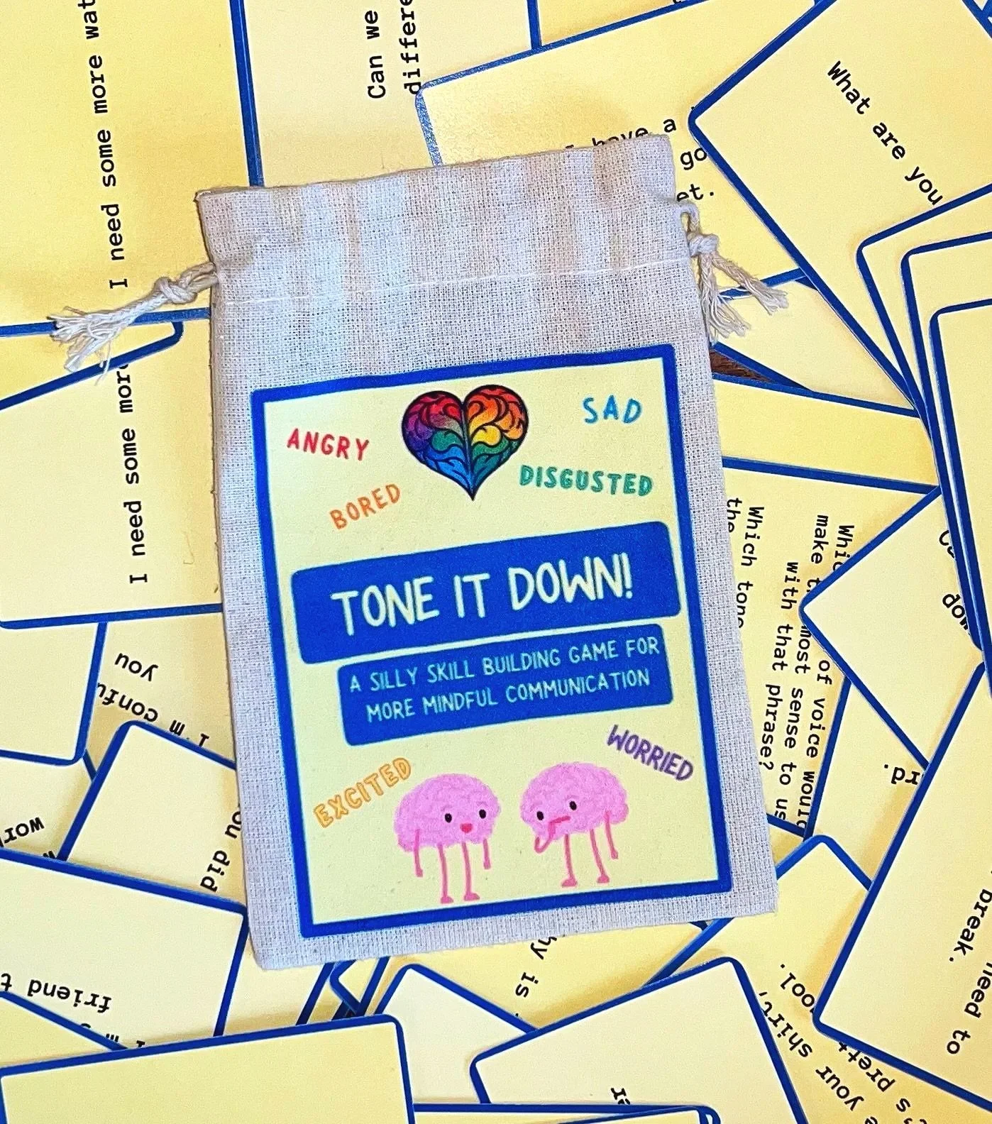 The image shows a card game called "Tone It Down" with a focusing on emotions, featuring a colorful heart with emotional words such as angry, sad, bored, disgusted, excited, worried. There are also cute illustrated pink characters with cheerful or worried expressions at the bottom of the card.