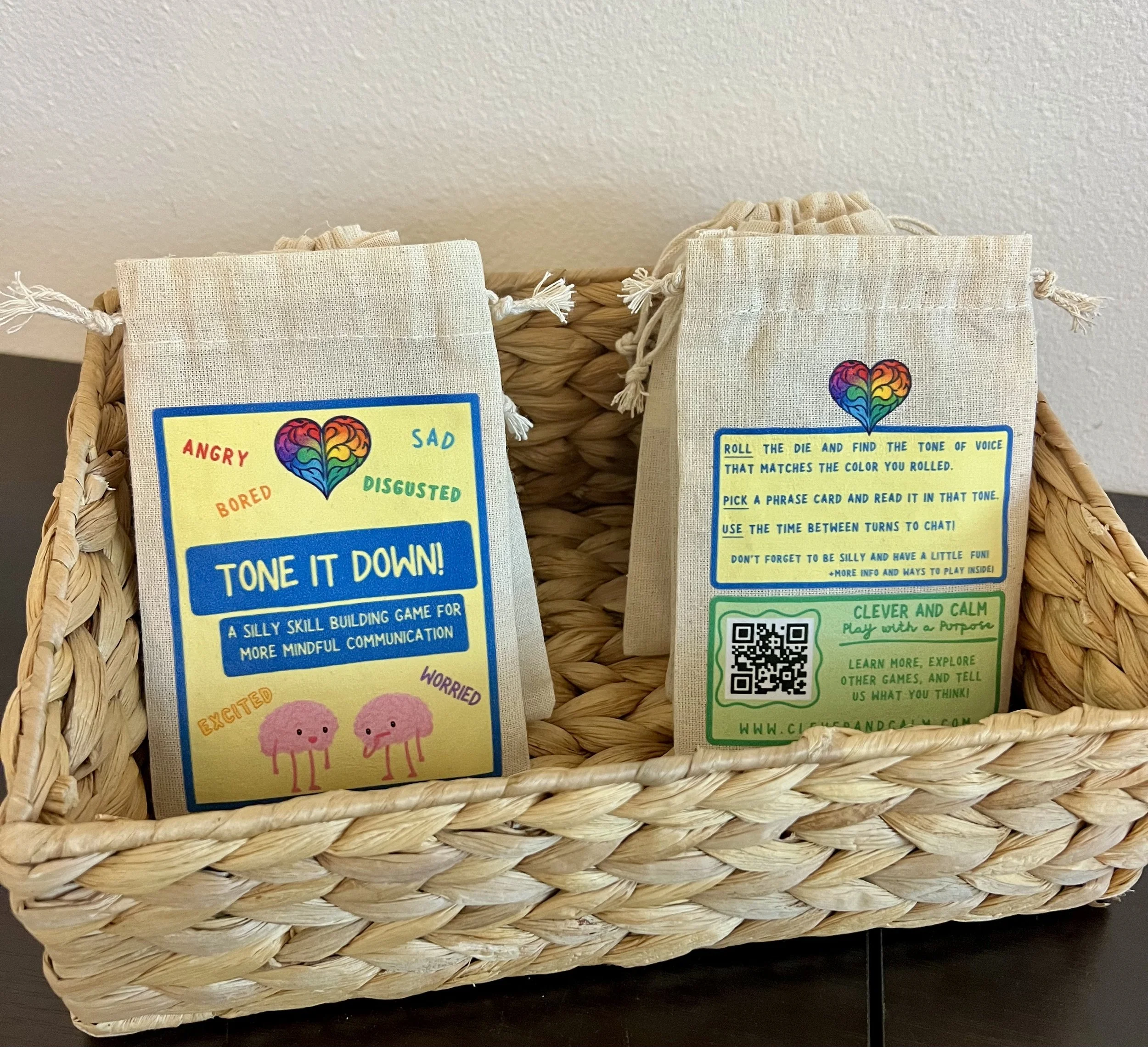 A wicker basket containing two small cloth pouches with educational game instructions, one with colorful emoji hearts representing emotions and another with a QR code and play tips, placed on a black surface against a plain wall.