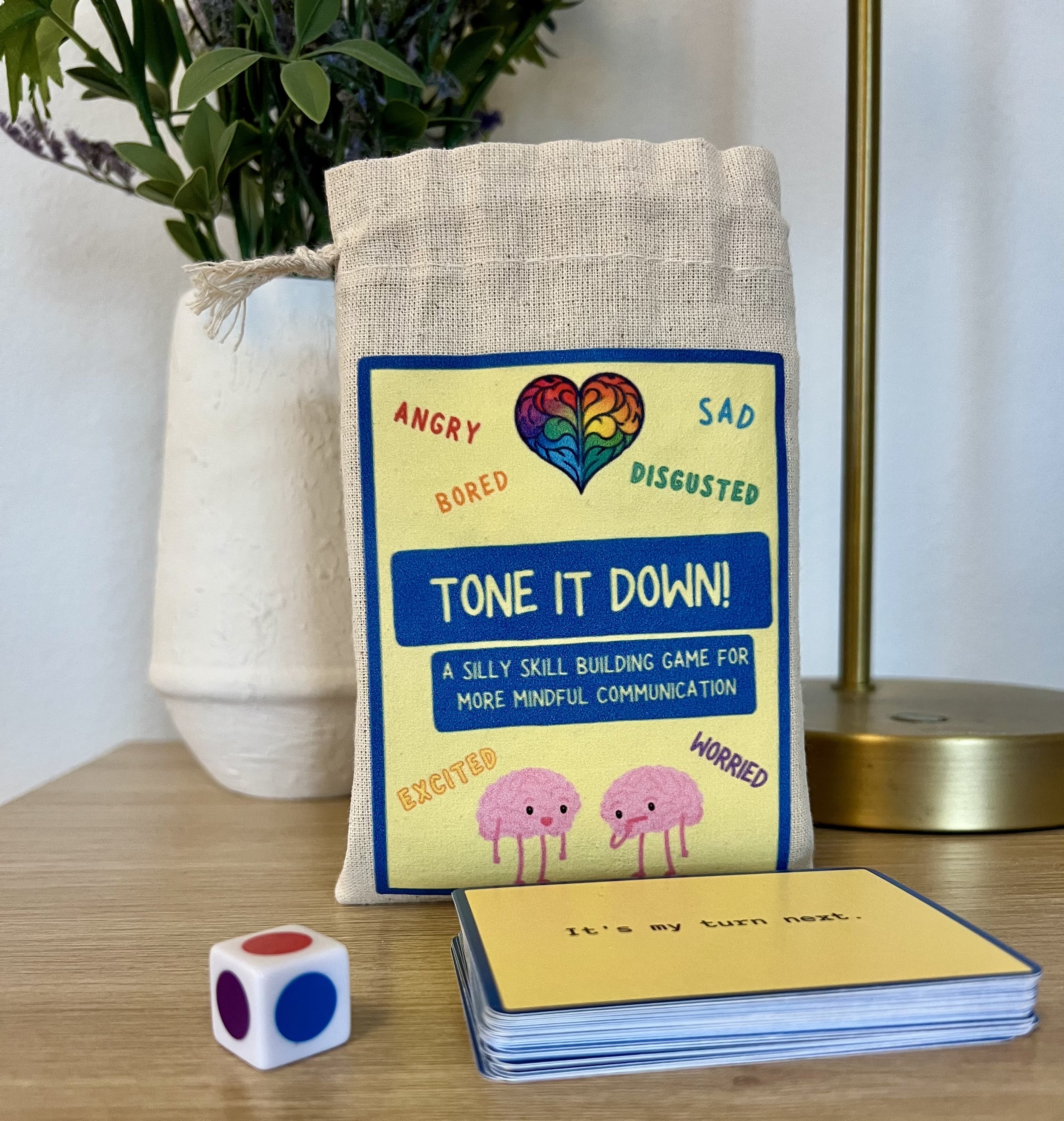 A game called 'Tone It Down' that helps children manage emotions, featuring characters representing different feelings such as angry, sad, bored, disgusted, excited, and worried. The game includes a colorful box, a dice, and a card with instructions, placed on a wooden table with a potted plant and a gold-colored lamp in the background.