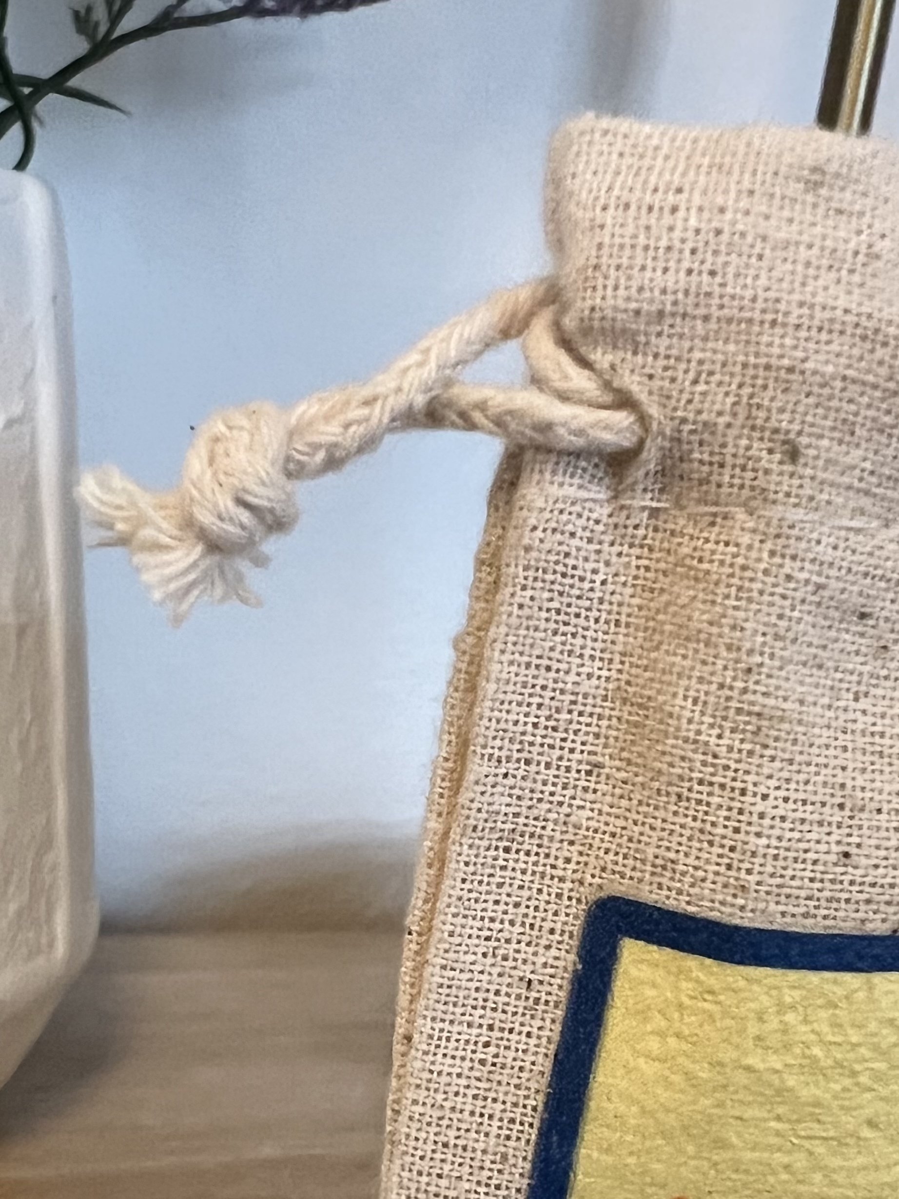 Close-up of a beige burlap bag with a twisted rope handle, placed on a beige woven mat with a blue and yellow rug next to it. Part of a plant and a piece of furniture are visible in the background.