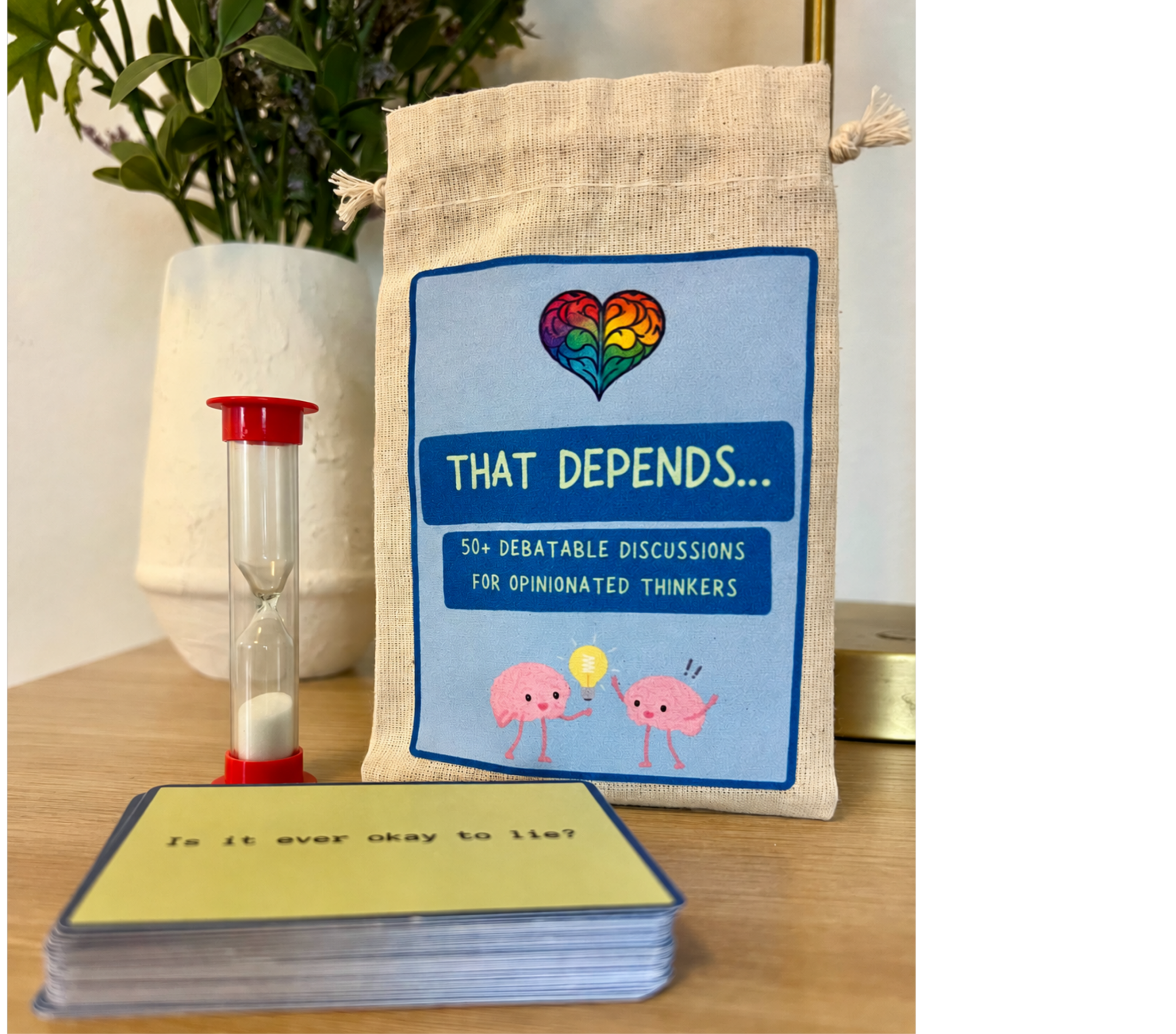A stack of several cards with a question visible on the top card: "Is it ever okay to lie?". Behind the cards, there is a beige tote bag with a blue label that has a colorful heart illustration and text about debatable discussions. To the left, there is a small clear hourglass with red ends, and in the background, a white vase with green plants and purple flowers.