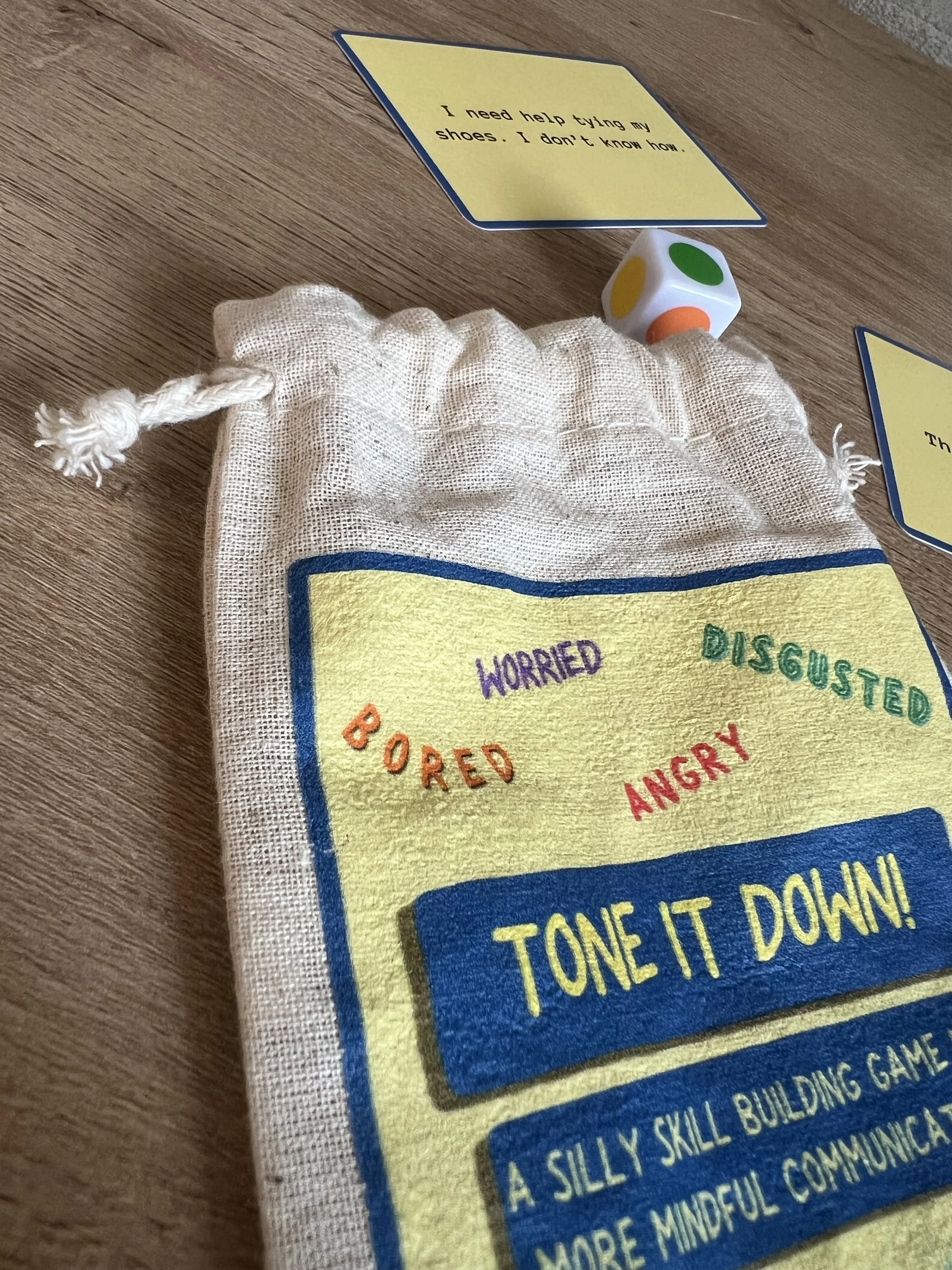 A small white bag with a colorful illustration of a board game and a die with green, yellow, and orange dots. The bag has a blue border and is partially visible on a wooden surface, with several yellow cards nearby. One card reads, 'I need help tying my shoes. I don't know how.'