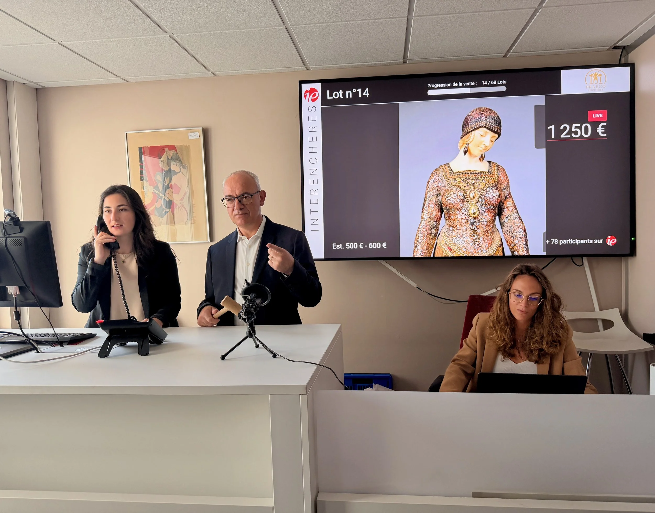 Une salle d'enchères où deux personnes sont visibles, une femme parlant au téléphone et un homme qui parle ou donne une explication, avec un grand écran derrière eux montrant un objet, un vêtement vestimentaire en mosaïque, avec un prix de vente affiché à 1250 euros. Il y a aussi d'autres personnes présentes, notamment une femme avec des lunettes assise à une table.