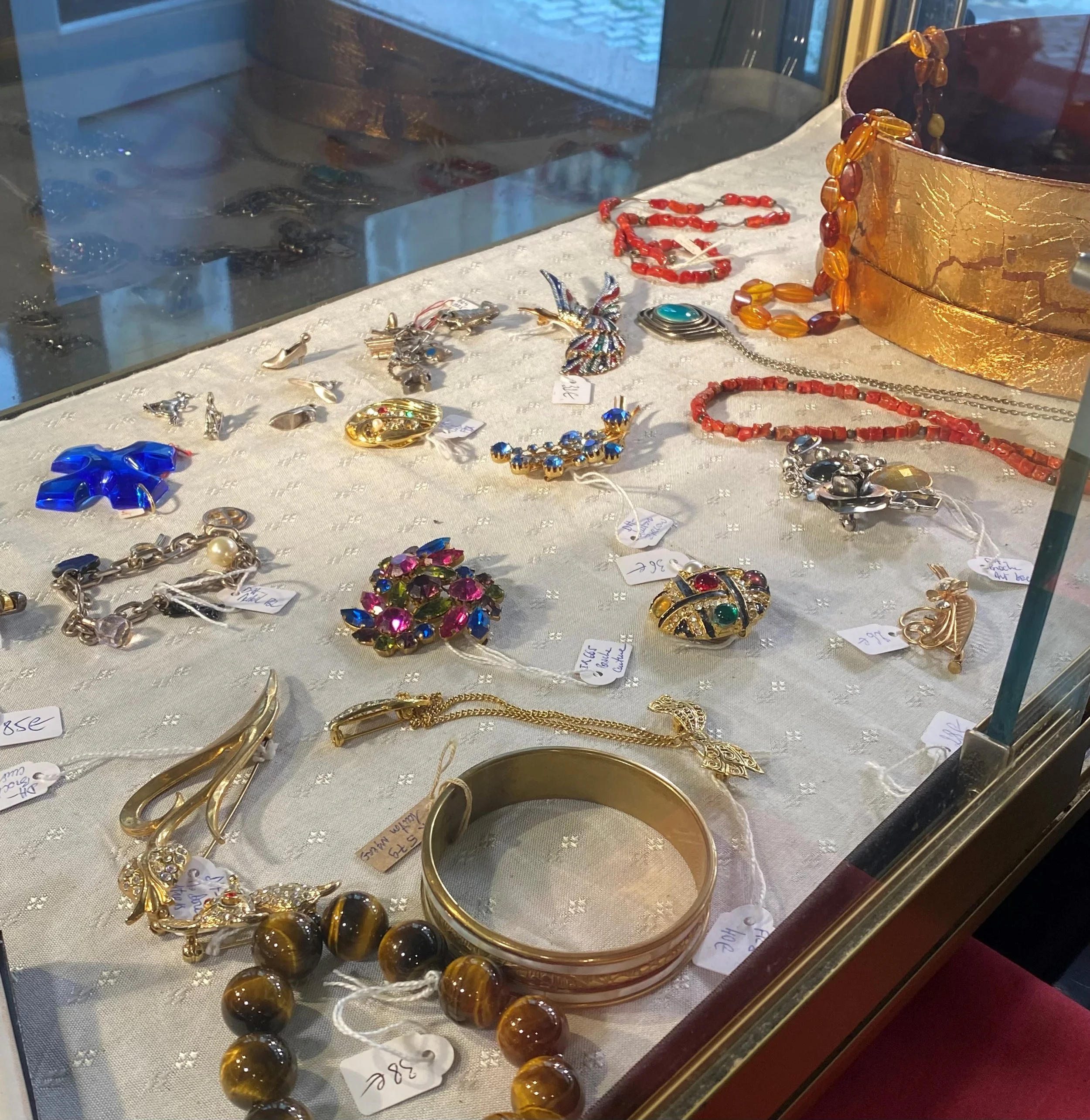 Vitrine d'objets en bijouterie vintage, comprenant des colliers, des bracelets et des broches aux couleurs vives, avec des étiquettes de prix et un fond sombre.