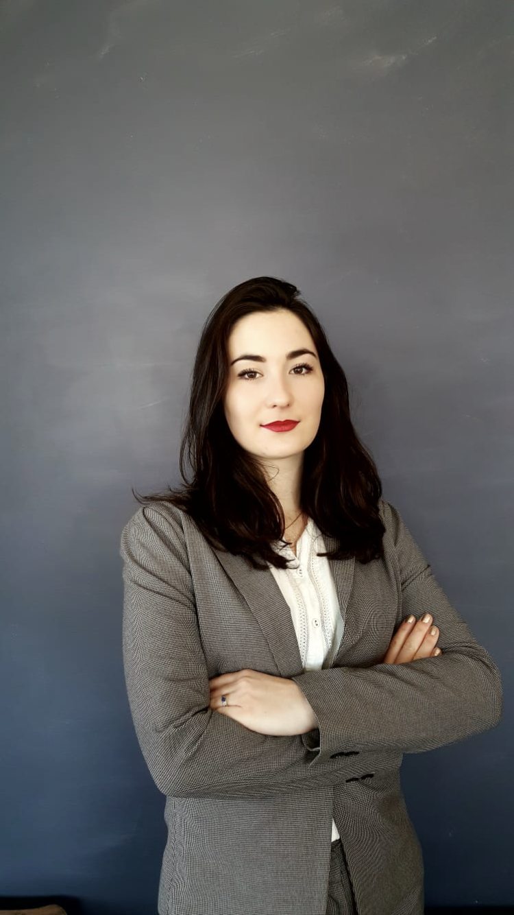 Une femme en costume gris foncé avec une chemise blanche, les bras croisés, devant un mur gris.