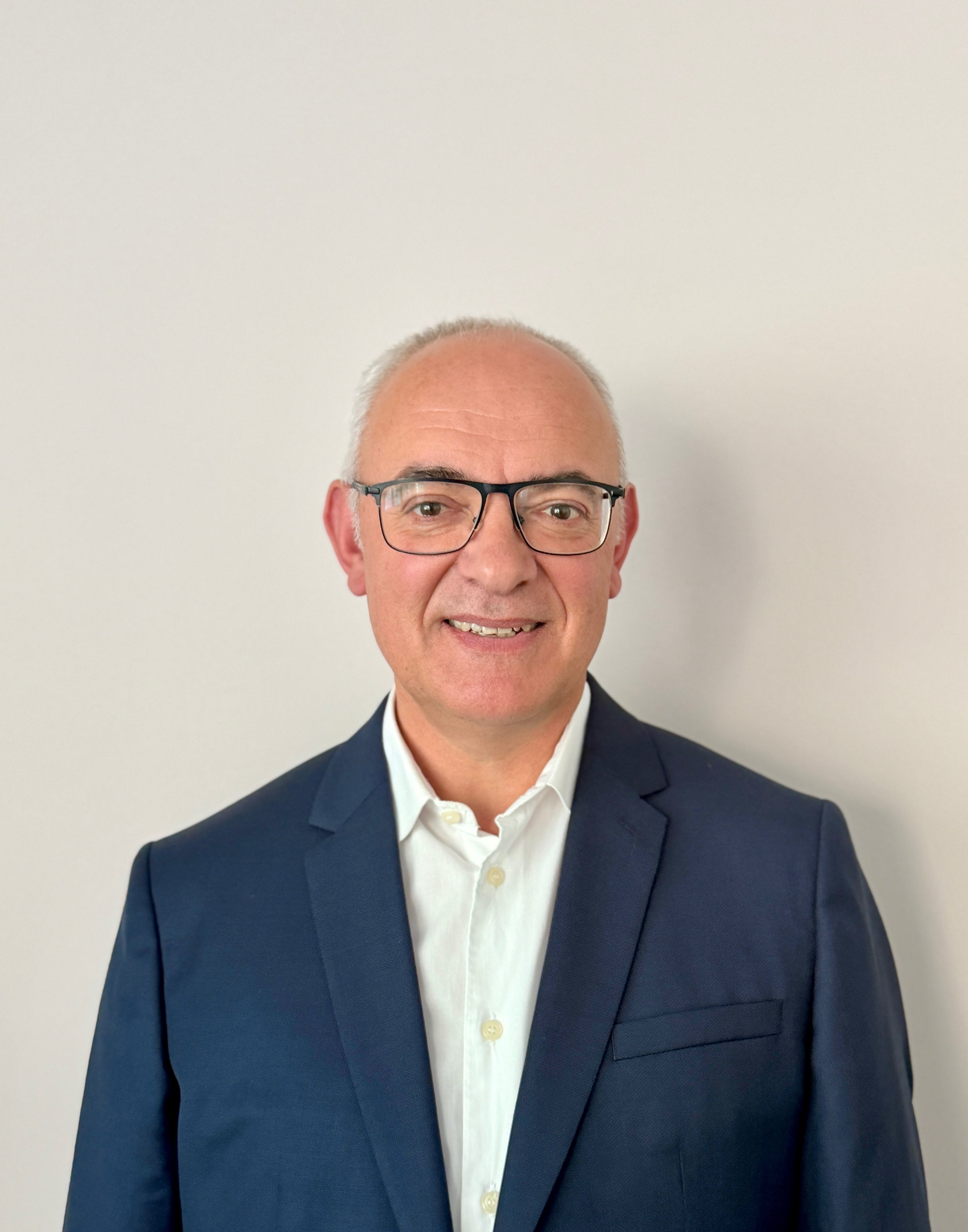 Portrait d'un homme d'affaires portant un costume bleu, une chemise blanche et des lunettes, souriant devant un mur blanc.
