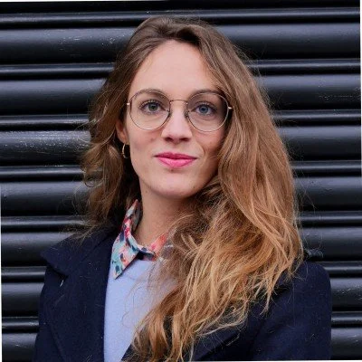 Jeune femme avec lunettes, cheveux ondulés, portant une veste sombre et chemise à col, posant devant un mur en métal noir.