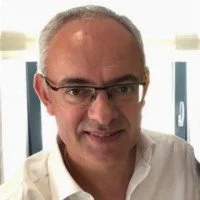 Homme avec des lunettes, cheveux gris, portant une chemise blanche, souriant, dans un intérieur lumineux.