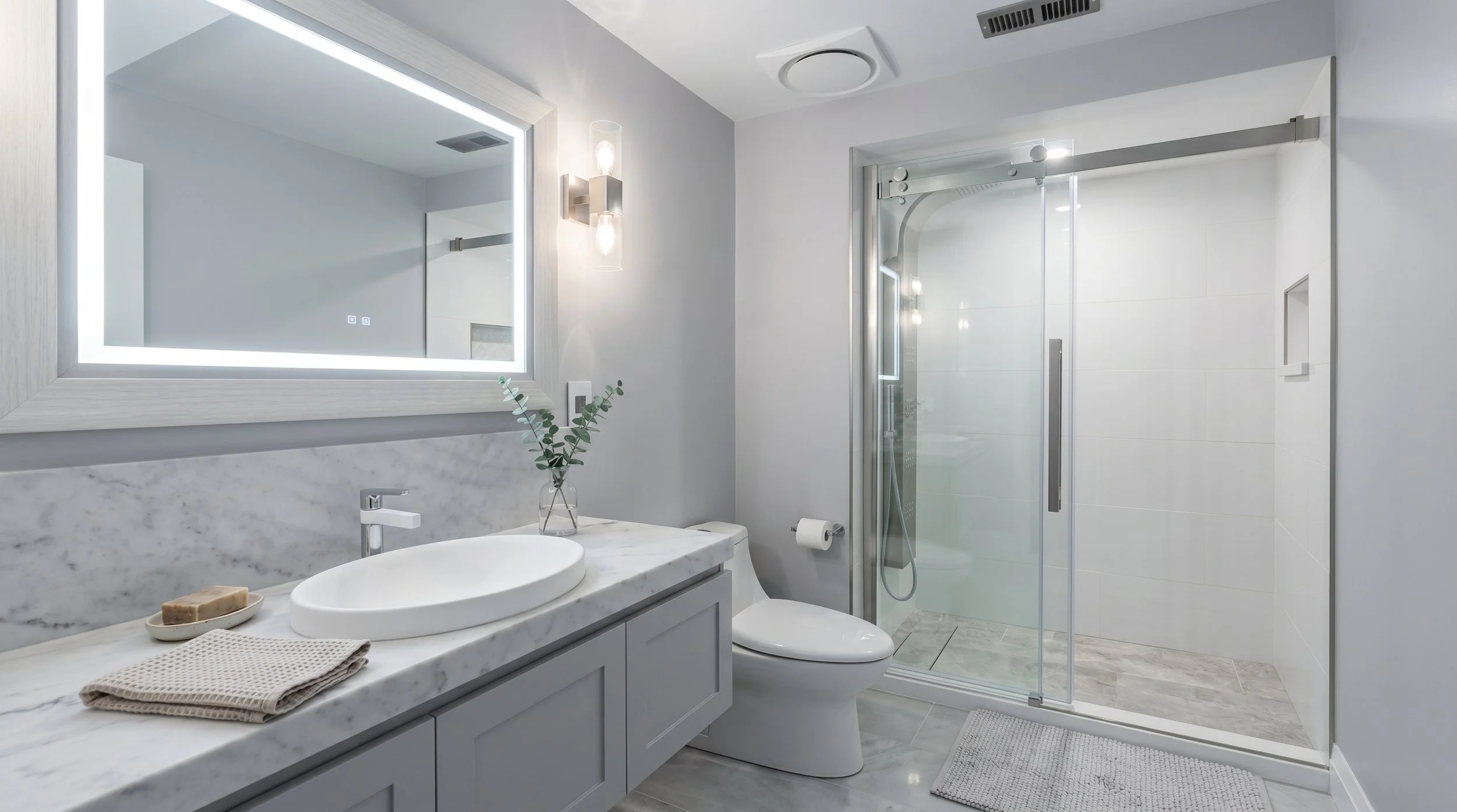 A modern bathroom with a large illuminated mirror above a white marble countertop sink, a glass shower enclosure, and minimalist decor.