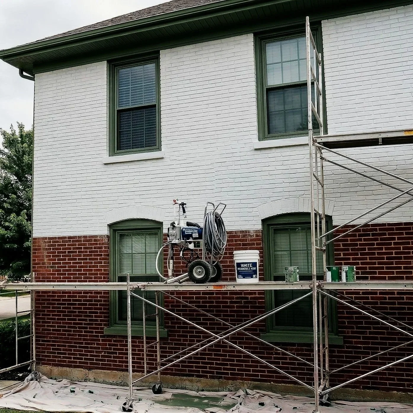 Construction scaffolding set up outside a building with white and red brick walls, green window frames, and a painter's equipment and paint bucket on the scaffolding platform.