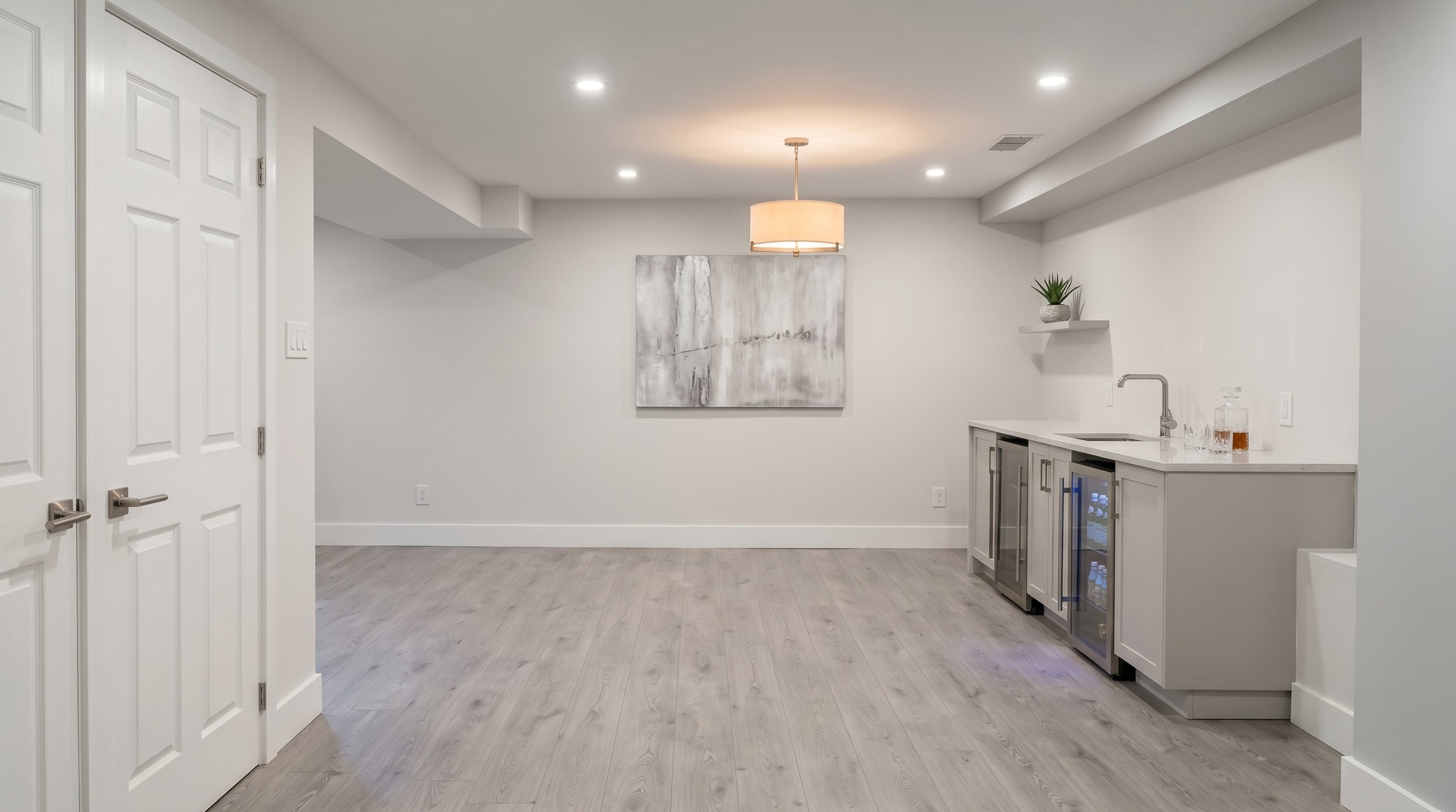 Interior of a modern basement with light grey walls, a small wet bar with a sink, glass barware, wine refrigerator, and a floating shelf with a potted plant. A large abstract painting hangs on the wall, and a ceiling light fixture provides ambient lighting.