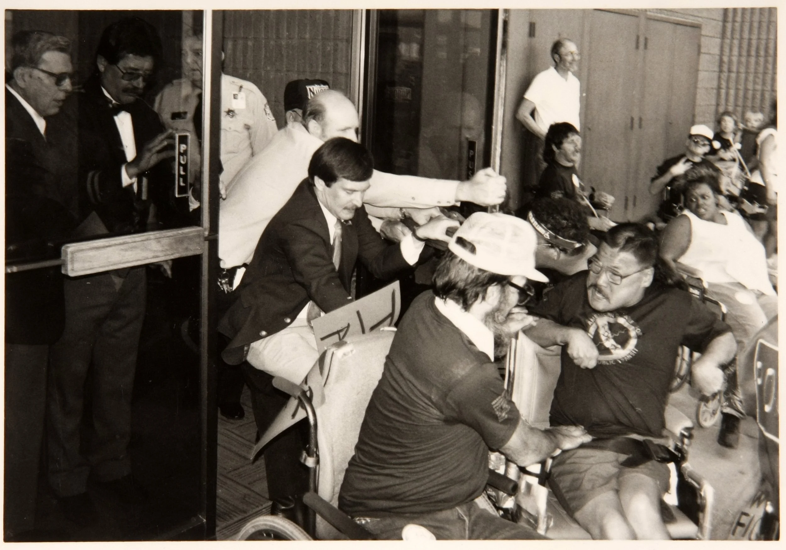 Archival photo of wheelchair users pressed against a building's glass doors while men in suits and uniformed officers hold the doors shut from inside
