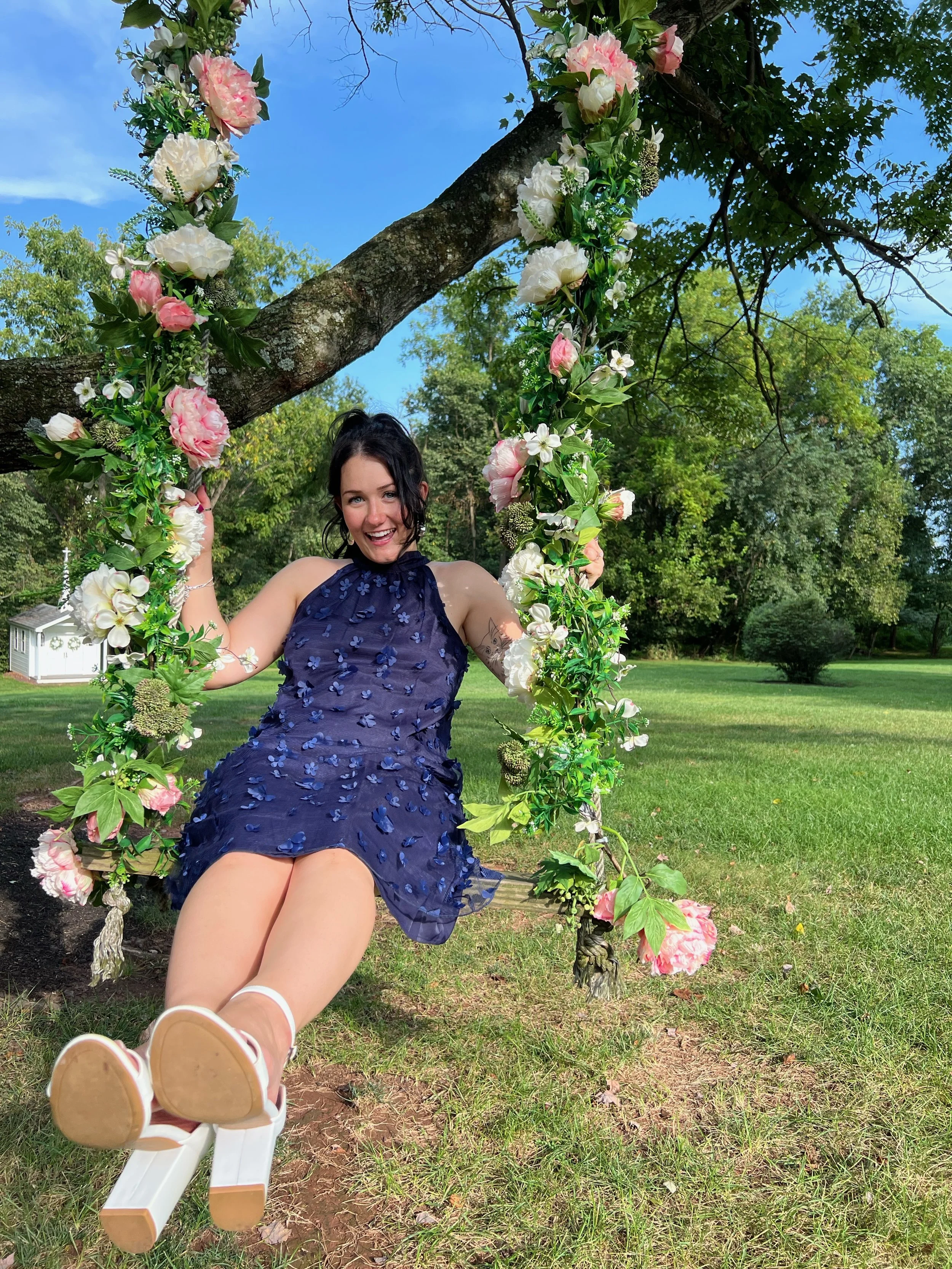 A woman sitting on a floral swing hanging from a tree, outdoors in a grassy park with trees and a blue sky in the background. She is smiling and wearing a dark blue dress with floral embellishments, and white sneakers.