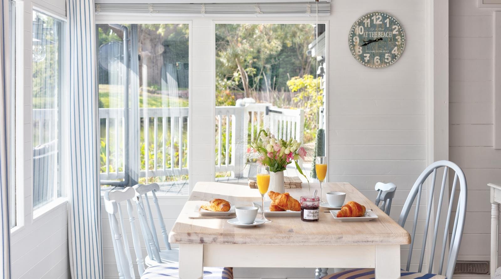 Bright breakfast nook with a wooden table set with croissants, coffee cups, two glasses of orange juice, a vase of flowers, and a jar of jam, next to a large window overlooking a porch and outdoor greenery, with a white wall clock showing 9:10.