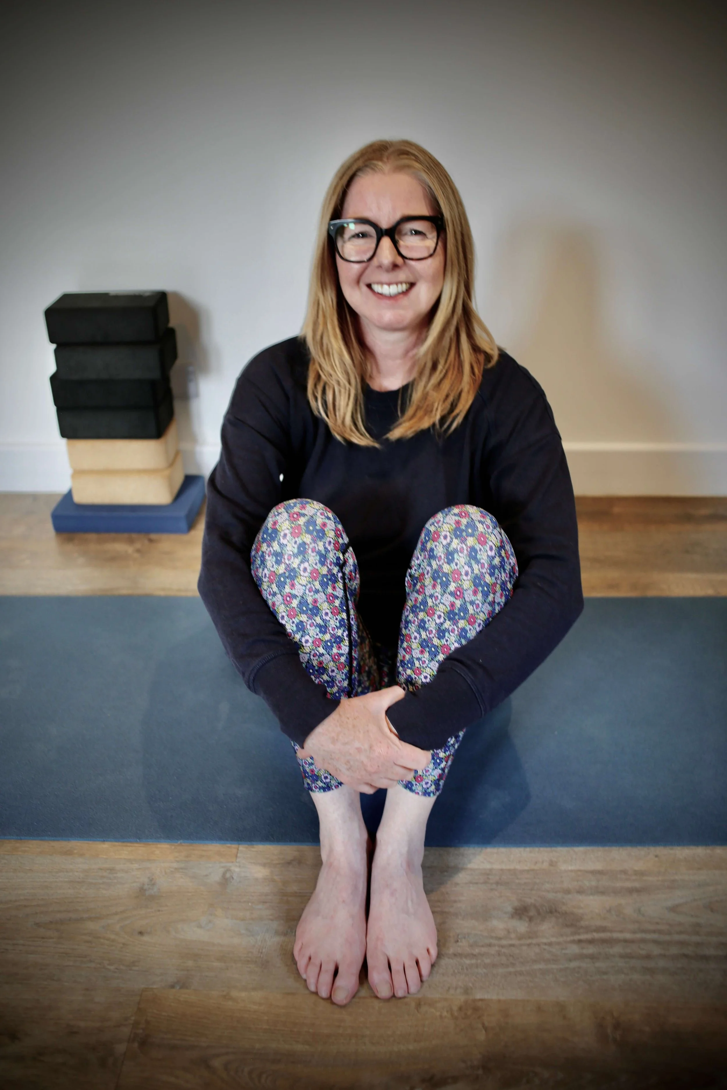 Catriona of Little Barn Yoga sitting cross-legged on a yoga mat indoors, smiling, wearing glasses, a black long-sleeve shirt, and floral-patterned leggings. There is a stack of foam blocks behind her on the floor and a plain white wall.