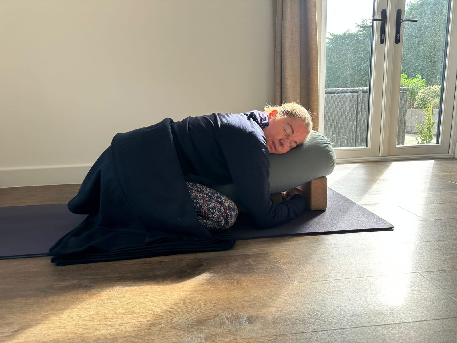 Catriona of Little Barn Yoga practicing yoga or meditation in a room with large windows. She is resting her head and arms on a bolster placed on a wooden platform, with her eyes closed and a peaceful expression.