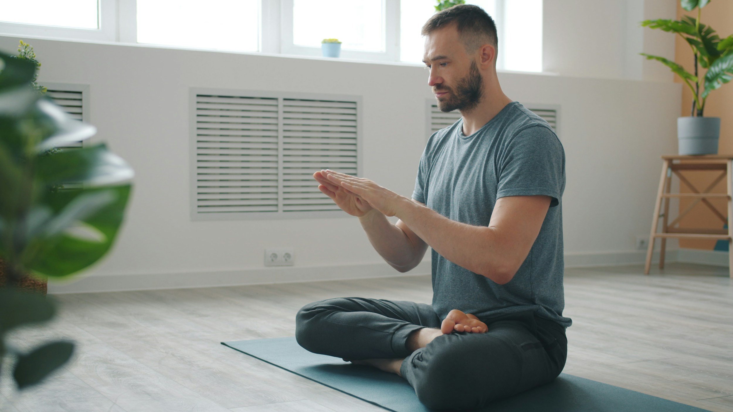 A man practicing yoga indoors, sitting cross-legged on a yoga mat with his hands in a prayer position, with natural light coming through the windows.