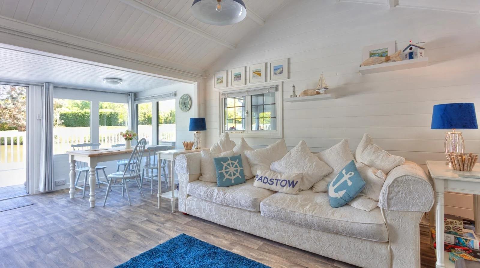 Bright seaside living room with white walls and ceiling, featuring a white sofa with nautical themed pillows, a window overlooking a yard with green trees, and a dining area with a white table and chairs near large windows.