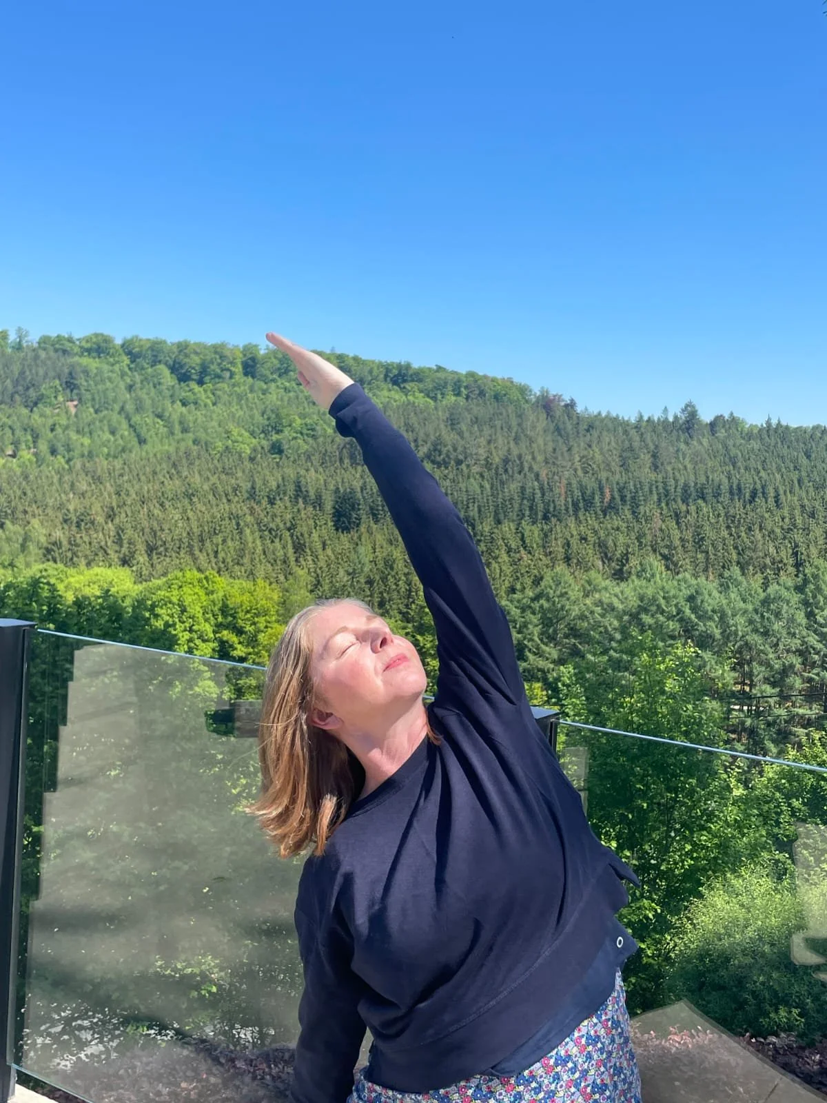 Catriona of Little Barn Yoga standing outdoors near a glass railing, stretching her arm upward, with her eyes closed against a backdrop of green trees and a clear blue sky.