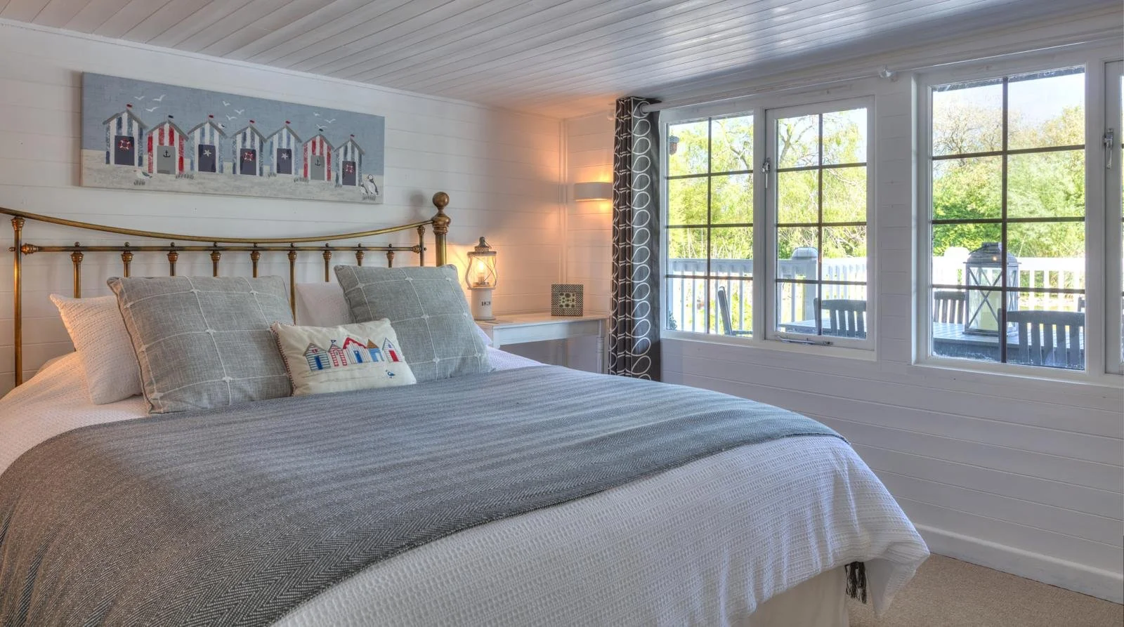 Cozy bedroom with large window, white wooden walls, and a bed with gray and white bedding, seagull artwork, and a pillow with beach hut embroidery.