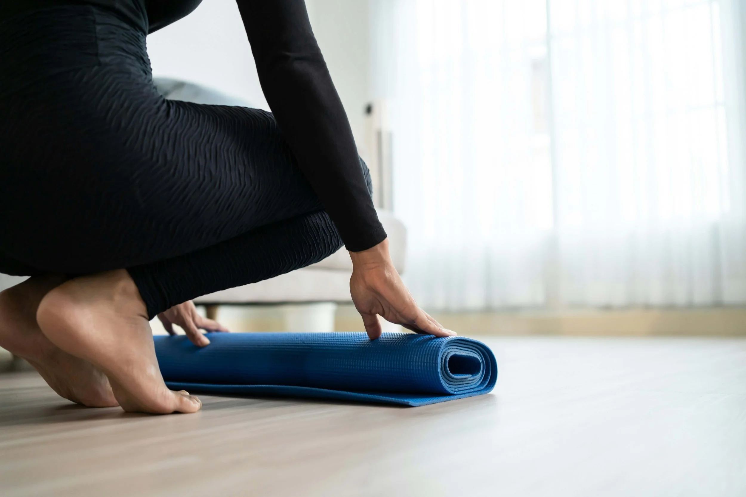 Person practicing yoga on a blue yoga mat indoors, focusing on their hand and part of their body, with a bright and minimalistic background.