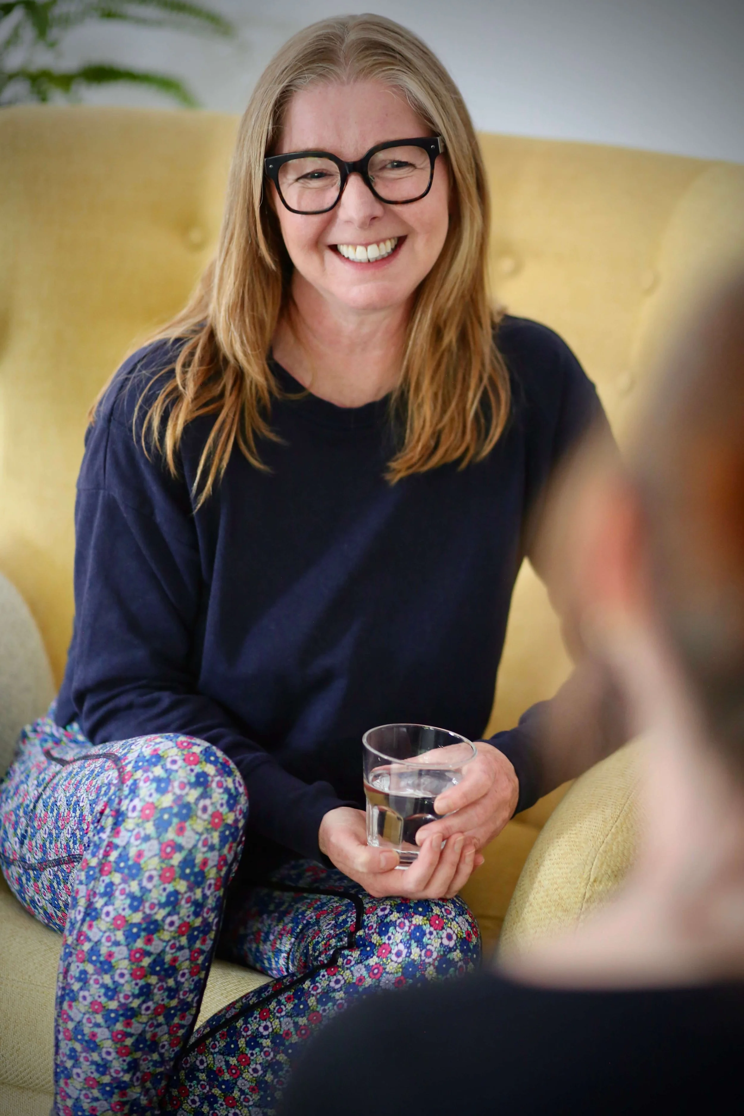 Catriona of Little Barn Yogawith long blonde hair, wearing glasses and a navy blue shirt, sitting on a yellow couch, holding a glass of water and smiling.