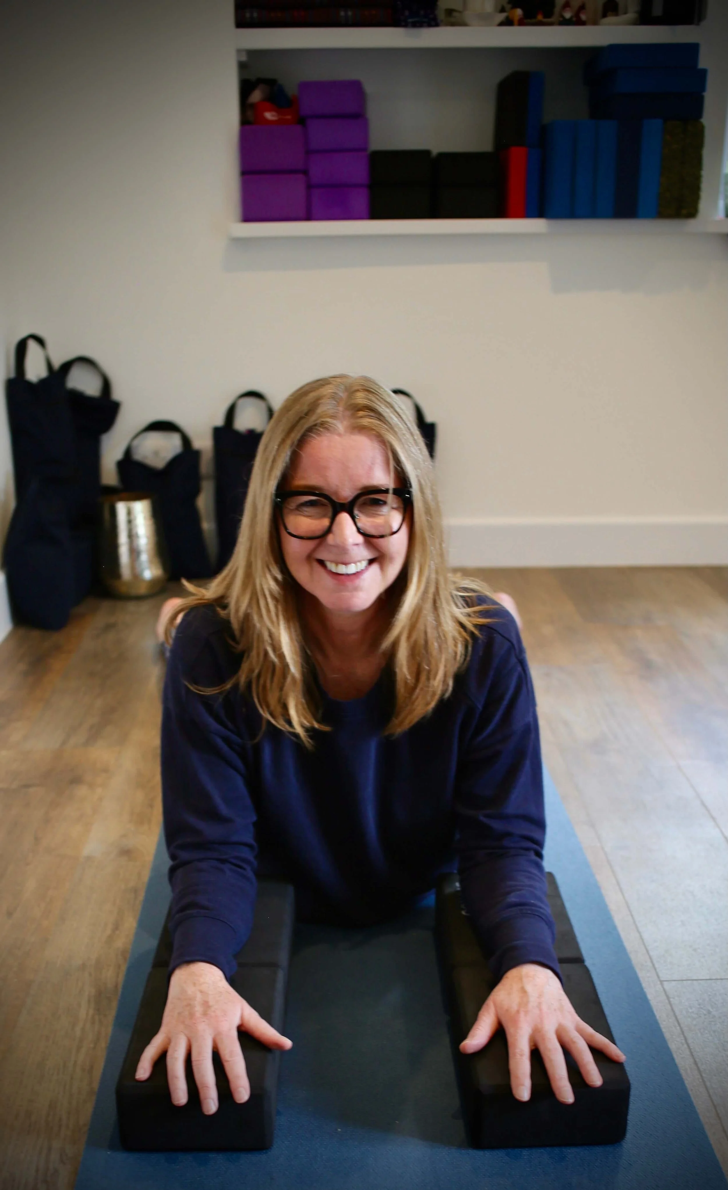 Catriona of Little Barn Yoga with glasses and long blonde hair smiling while practicing yoga on a black workout mat, with her hands resting on two black yoga blocks, in a room with a shelf of yoga blocks and bags in the background.