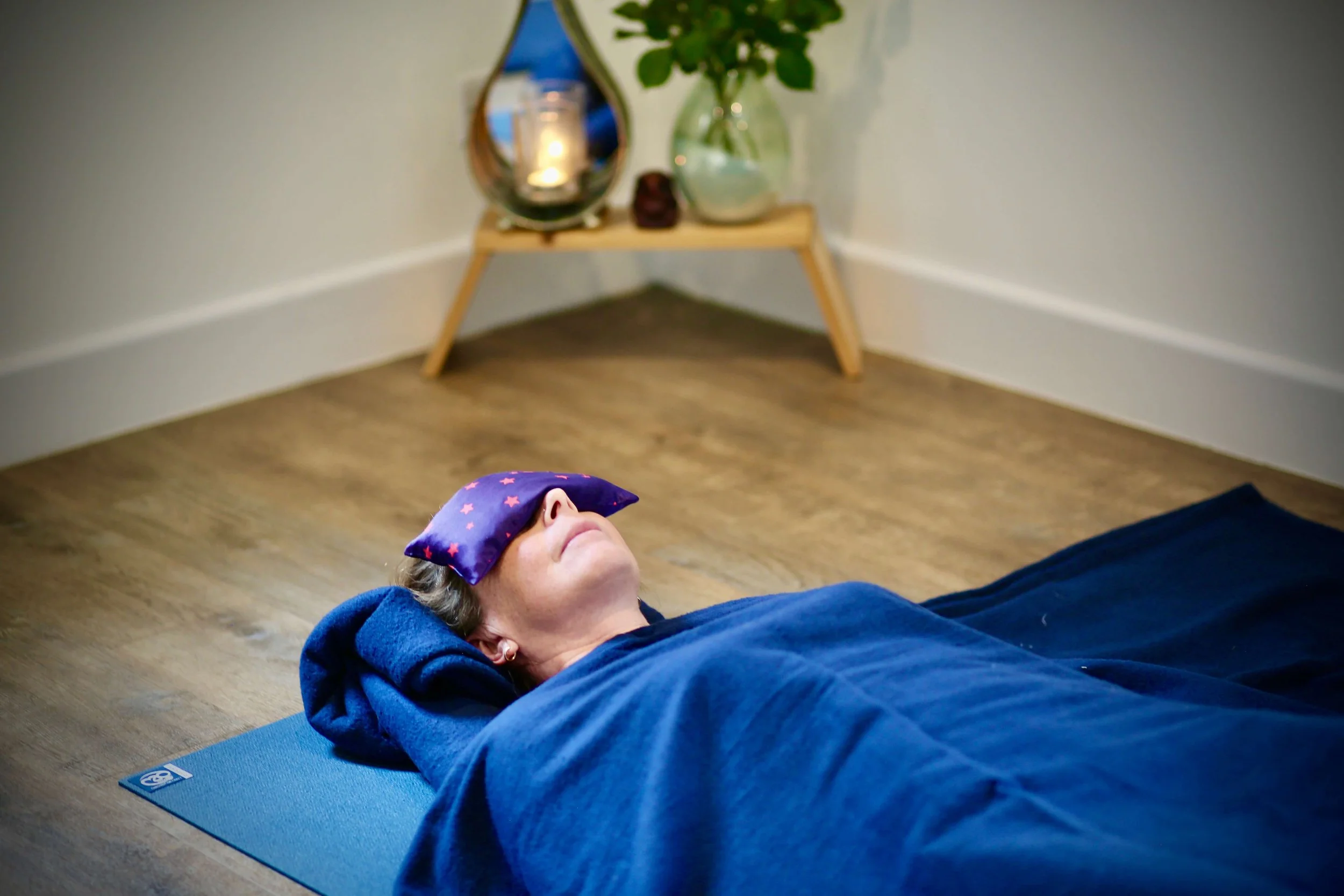A person lying on their back on a yoga mat, wearing a blue hoodie and a star-patterned sleep mask, in a room with light-colored walls and wooden flooring.