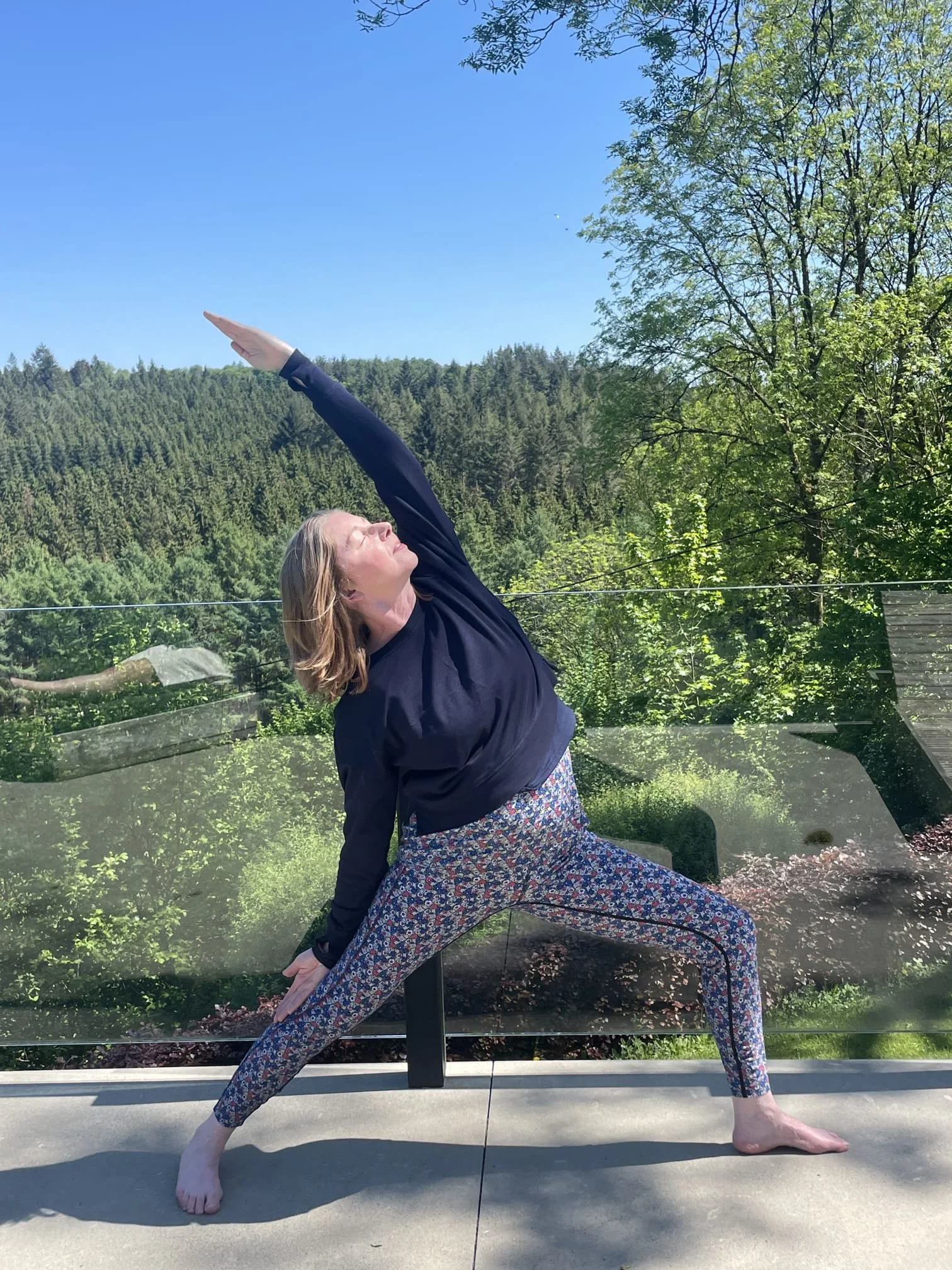 Catriona of Little Barn Yoga practicing yoga outdoors on a sunny day, performing a wide-legged stance with one arm reaching upward and the other arm extended downward, with a backdrop of green trees and a blue sky.