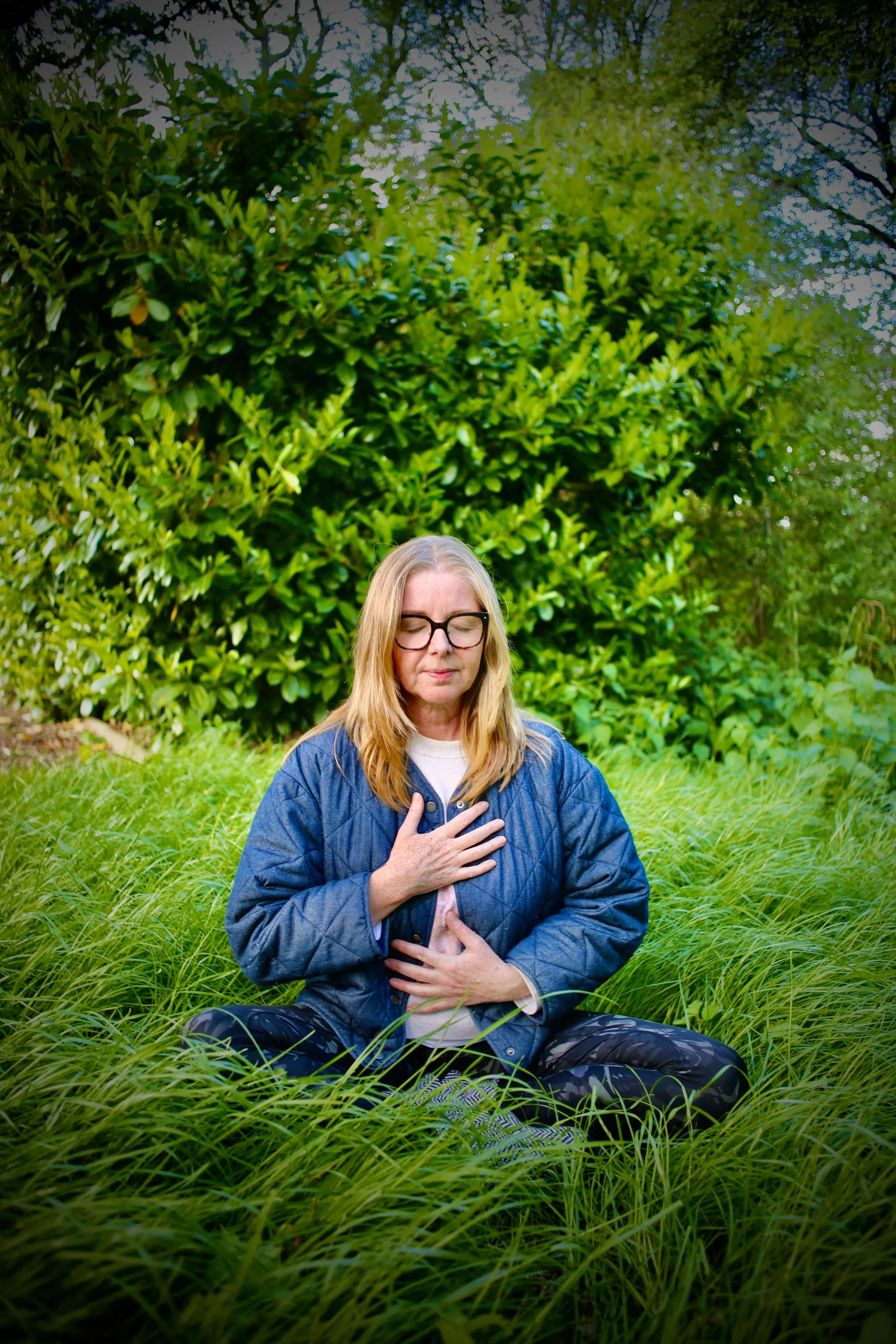 Catriona of Little Barn Yoga sitting cross-legged in a grassy field with her eyes closed, holding her chest and stomach, surrounded by green trees and plants, practicing meditation or mindfulness outdoors.