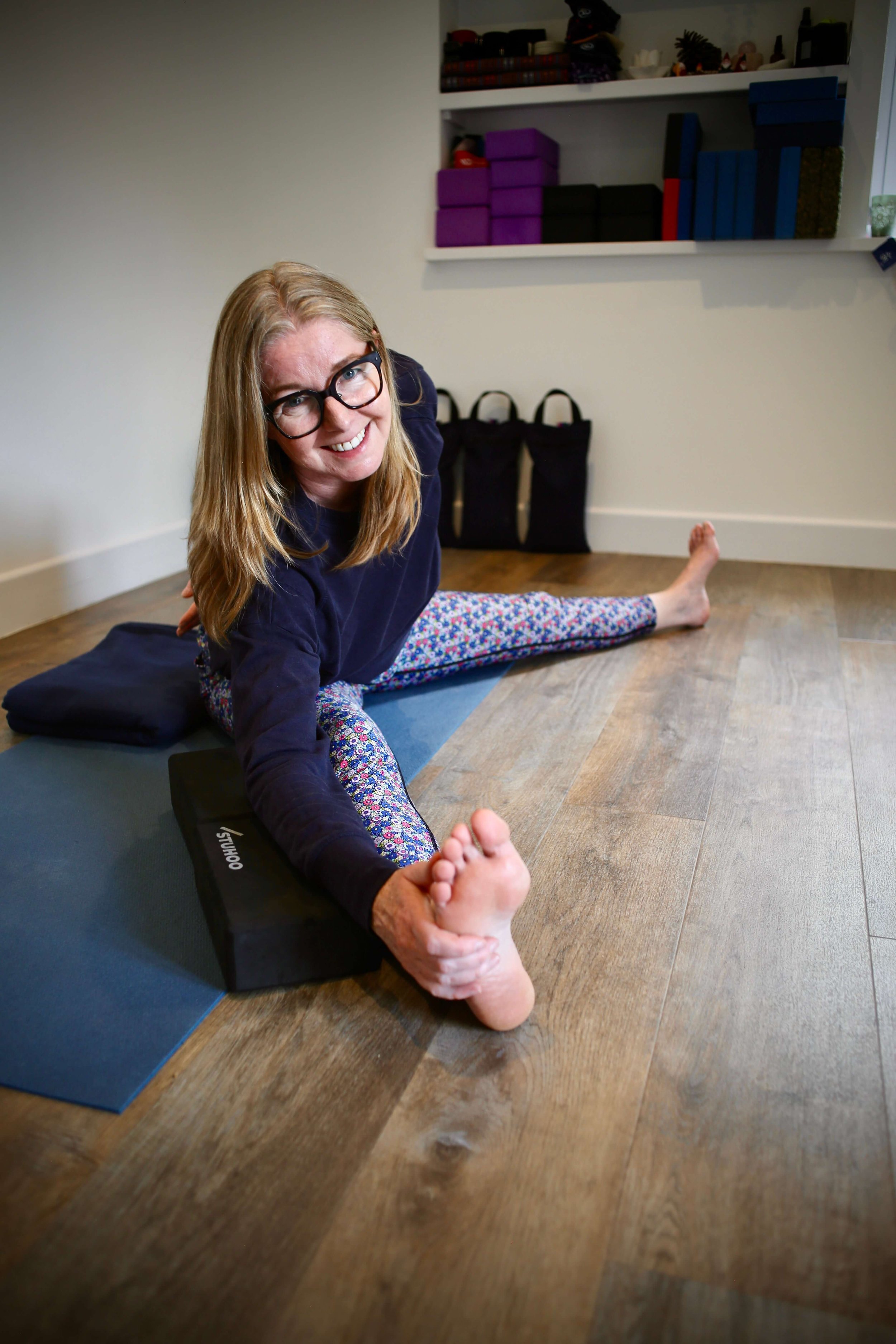 Woman performing a seated forward bend yoga pose on a mat indoors, smiling at the camera.