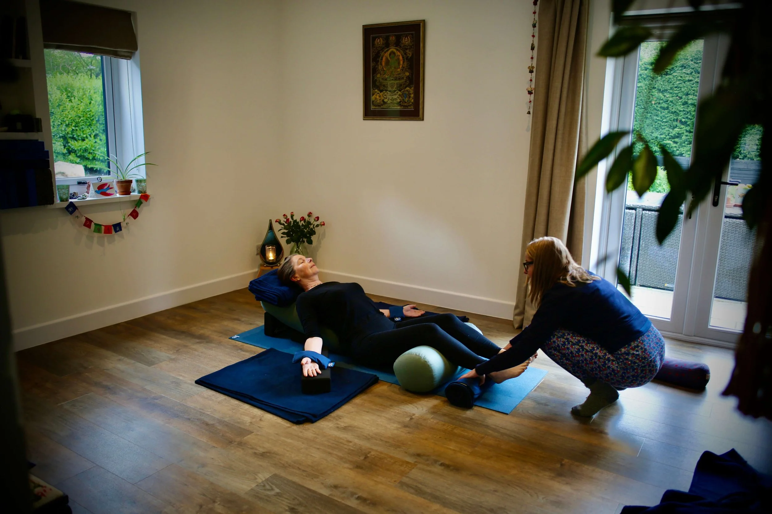 A person lying on a yoga mat with a pillow under their head, participating in a relaxation or meditation session. Another person is assisting them, and a third person is sitting nearby, possibly an instructor. The room is well-lit with natural light, decorated with plants, a framed picture on the wall, and a balcony door.