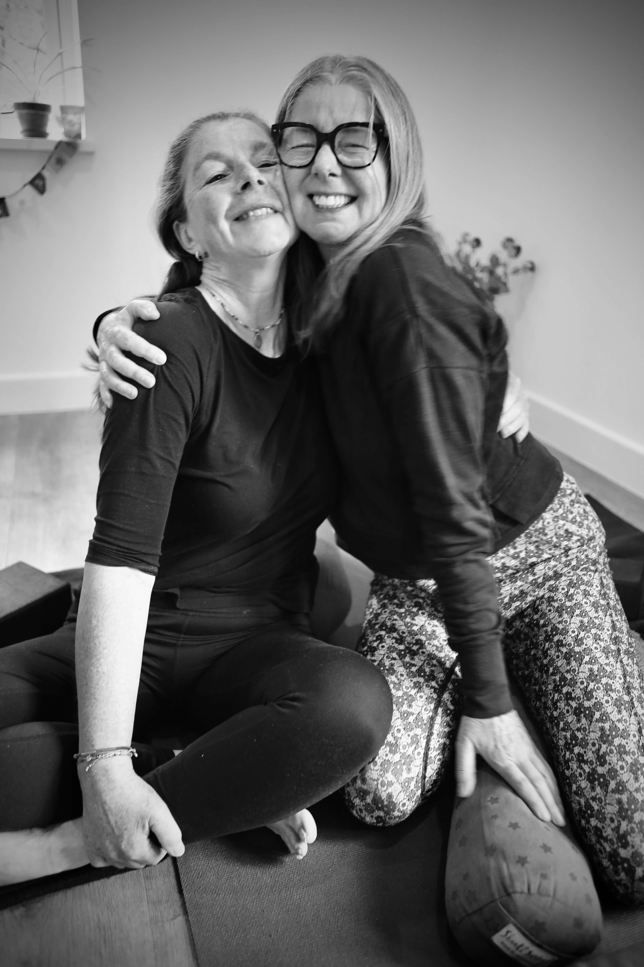 Two women hugging and smiling in a room. One woman is seated cross-legged, and the other woman is kneeling, with a ball and some objects beside them.