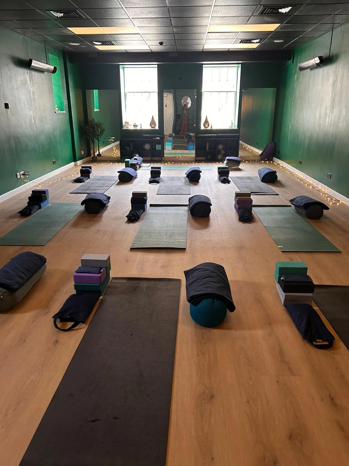 Yoga or meditation studio set up with yoga mats, bolsters, blocks, and blankets arranged on a wooden floor, with a green accent wall and large window.