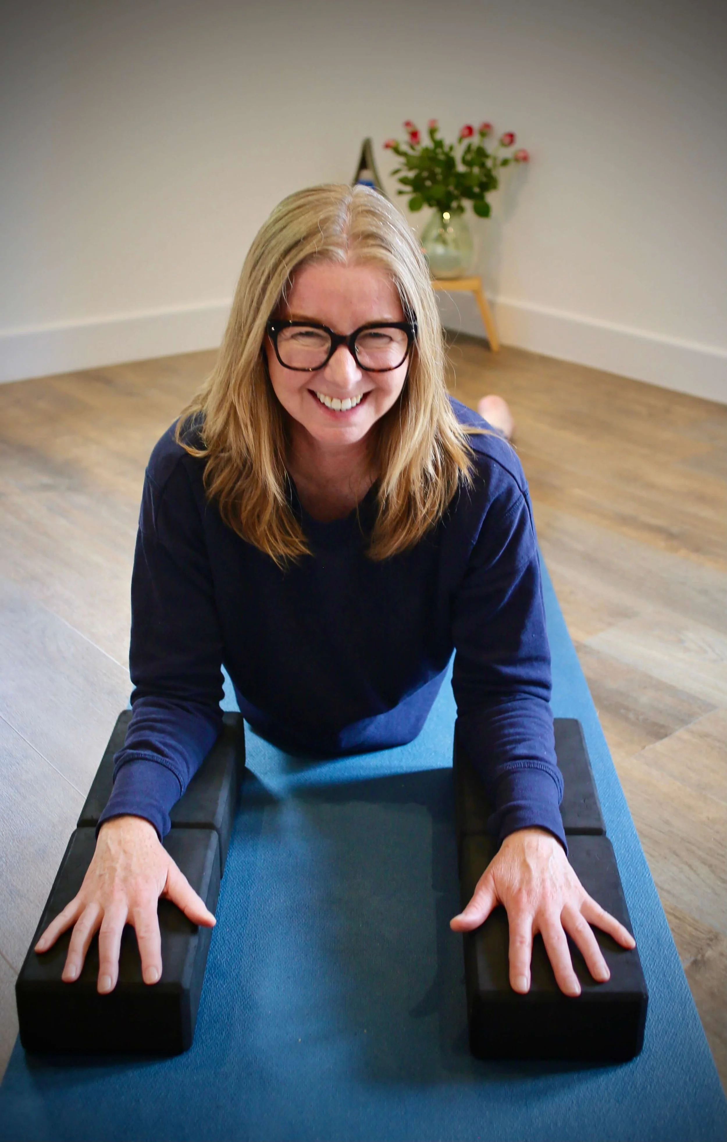 Catriona of Little Barn Yoga with long blonde hair, glasses, and a dark blue long-sleeve shirt in a well-lit room practicing yoga on a blue mat, with hands pressed on yoga blocks.
